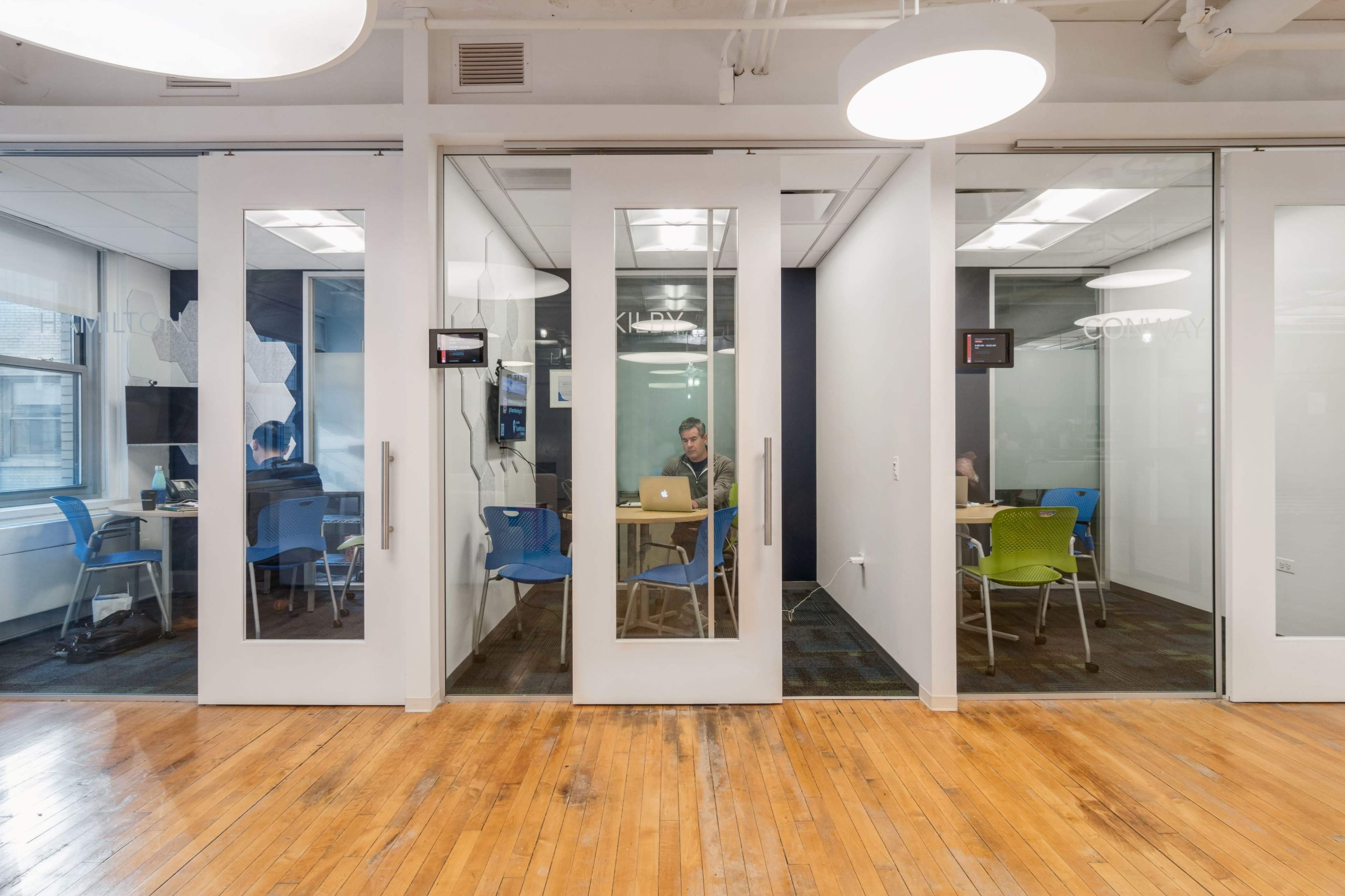 The image shows a modern office space with three transparent meeting rooms, each featuring a table and chairs, and one person working at a laptop inside the leftmost room.
