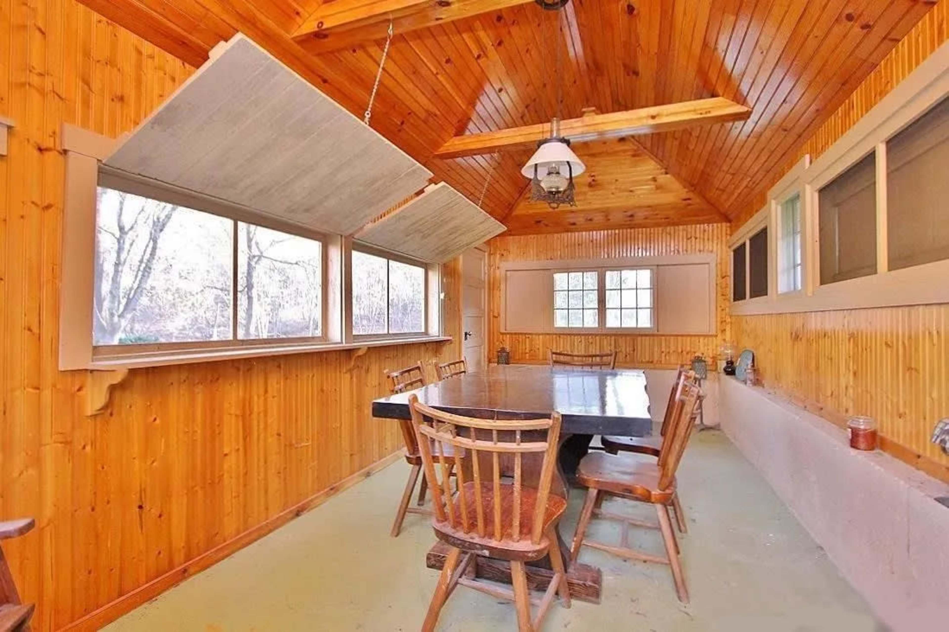 A wooden-paneled dining area with a large table and several chairs, alongside windows that open outward.