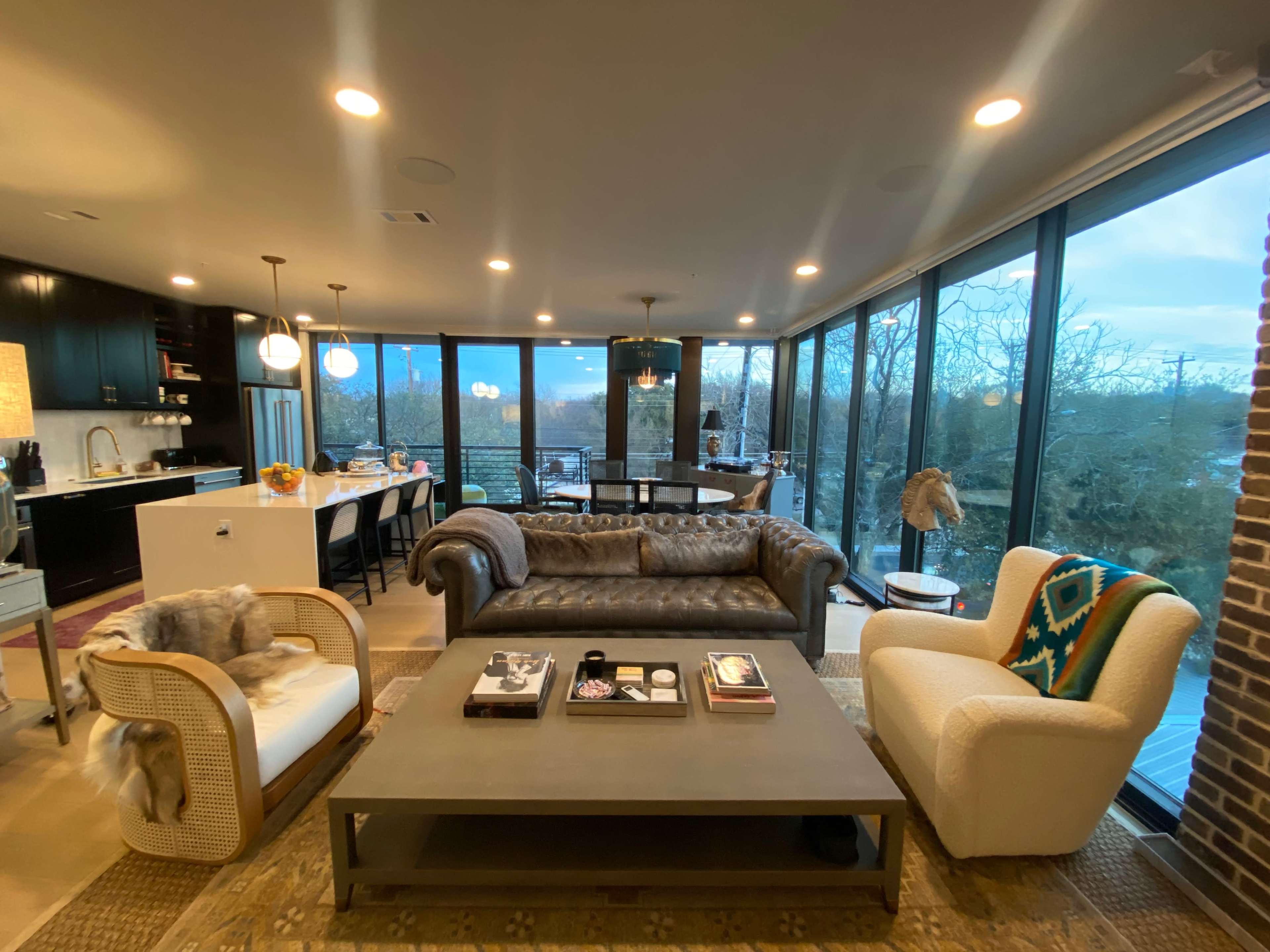 The image shows a modern living area with large windows, featuring a brown leather sofa, two armchairs, and a coffee table, all set in an open-concept layout that connects to a kitchen.