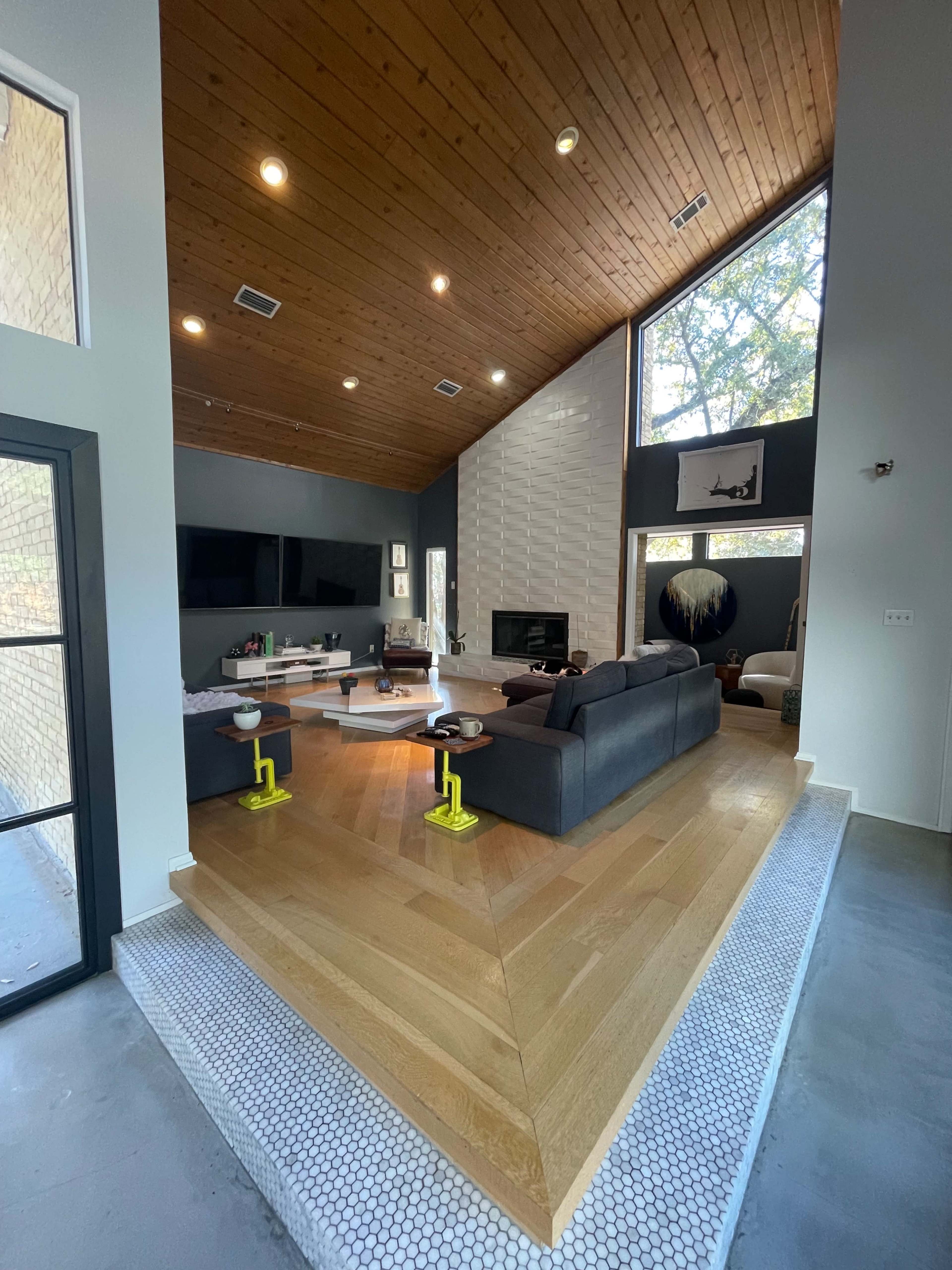 A modern living room with a high wooden ceiling, large windows, and a raised hardwood platform separating the seating area from the rest of the space.