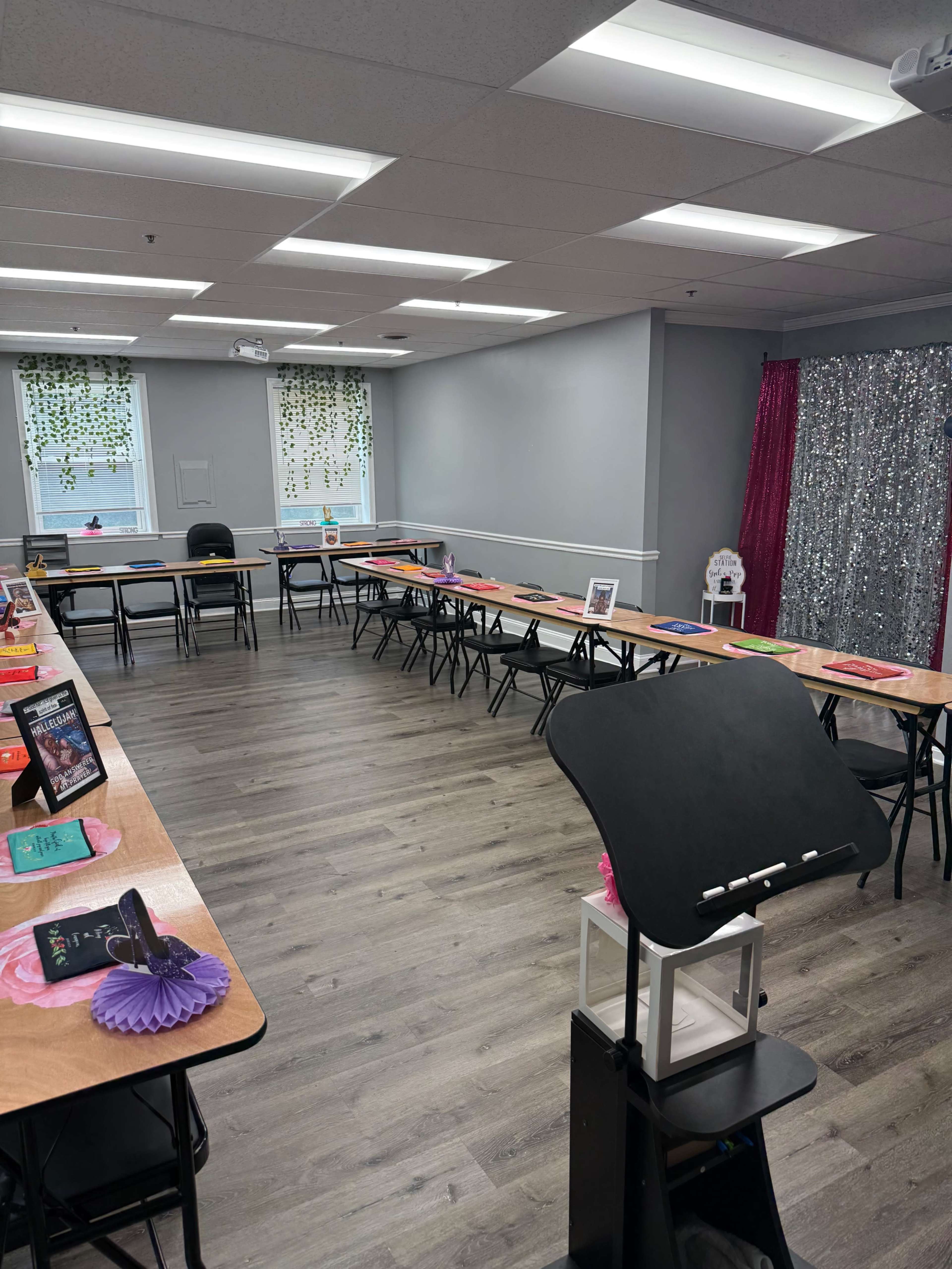 The image shows a classroom setup with multiple tables arranged in an open space, decorated with colorful materials and lighting, and a podium at the front.