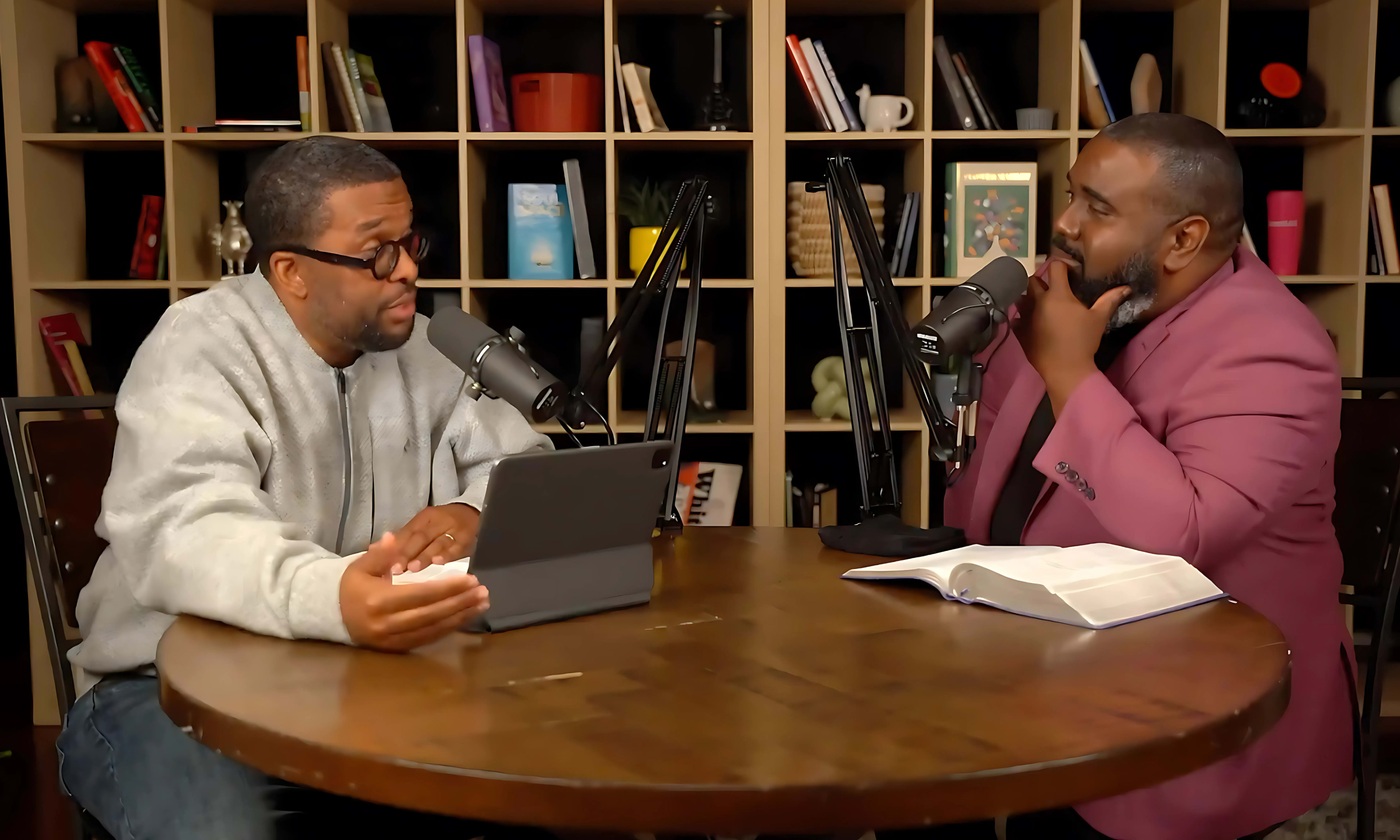 Two men are sitting at a round wooden table, engaged in a discussion, with microphones in front of them and a book open on the table.