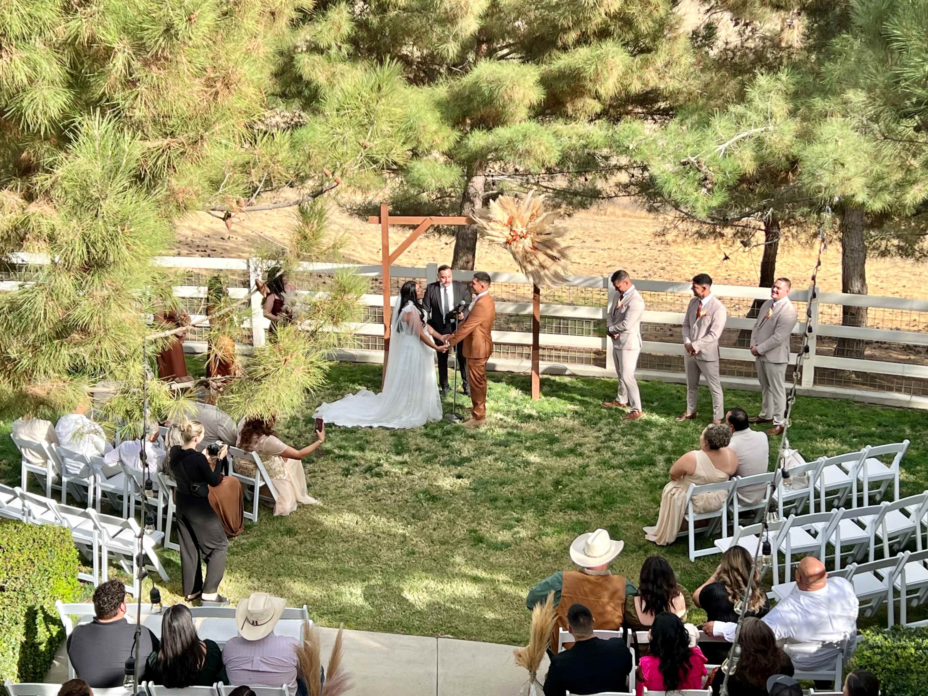 A couple is exchanging vows under a wooden arbor surrounded by guests seated on white chairs in a grassy outdoor setting.