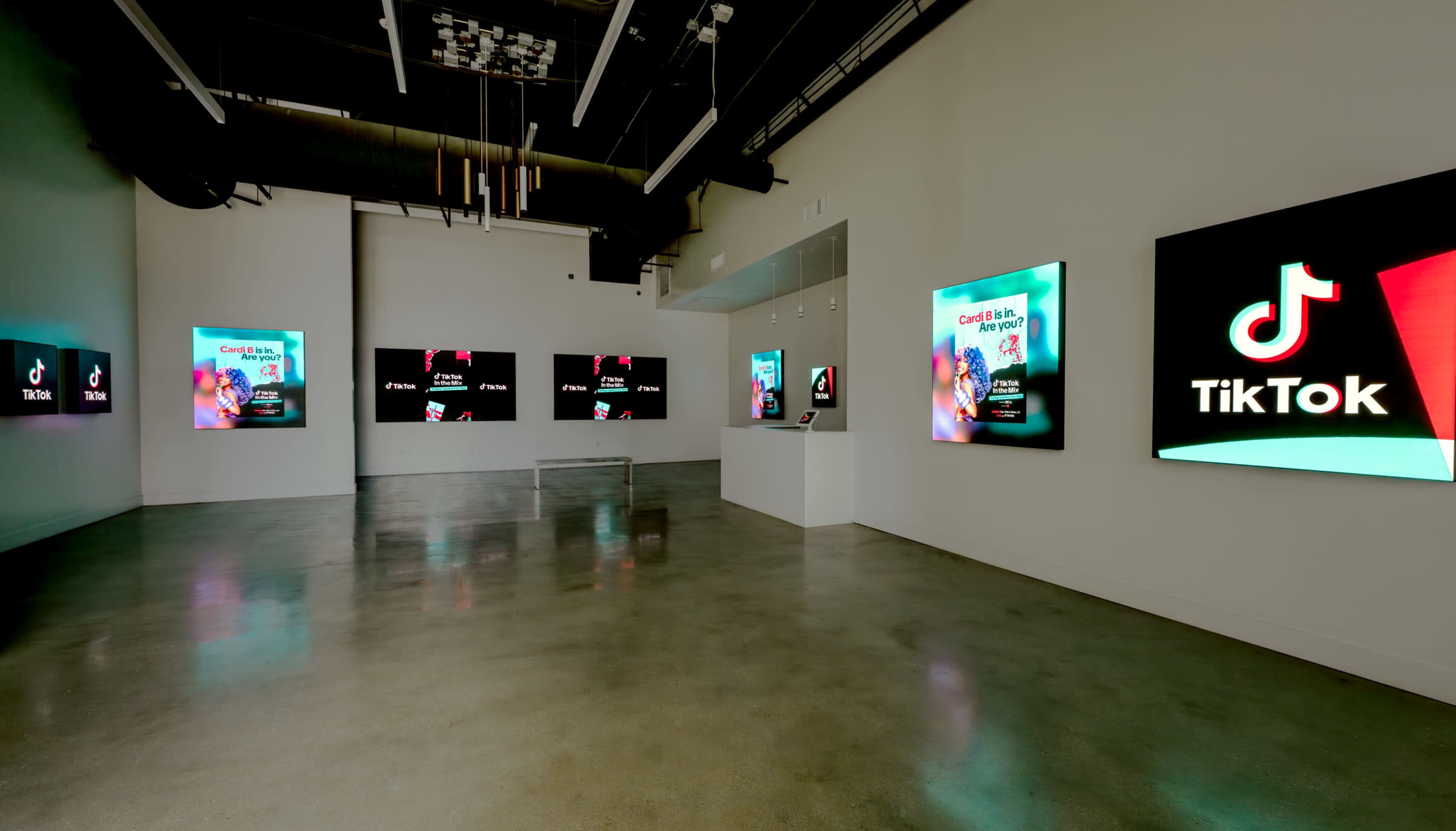 The image shows a modern exhibition space featuring multiple screens displaying TikTok content, with sleek polished flooring and minimalist lighting.