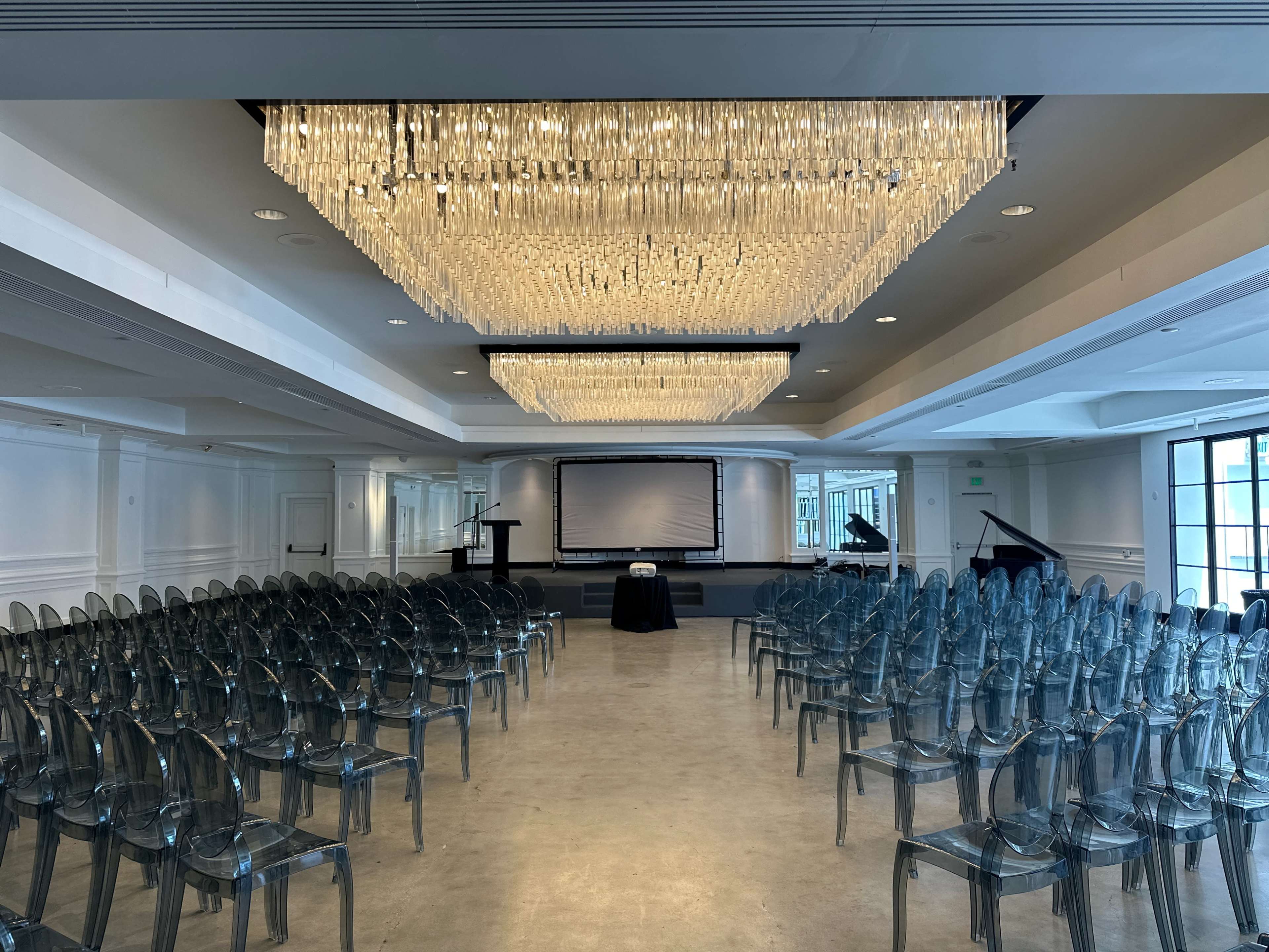 A spacious event room is arranged with rows of clear plastic chairs facing a presentation screen, under a large crystal chandelier.