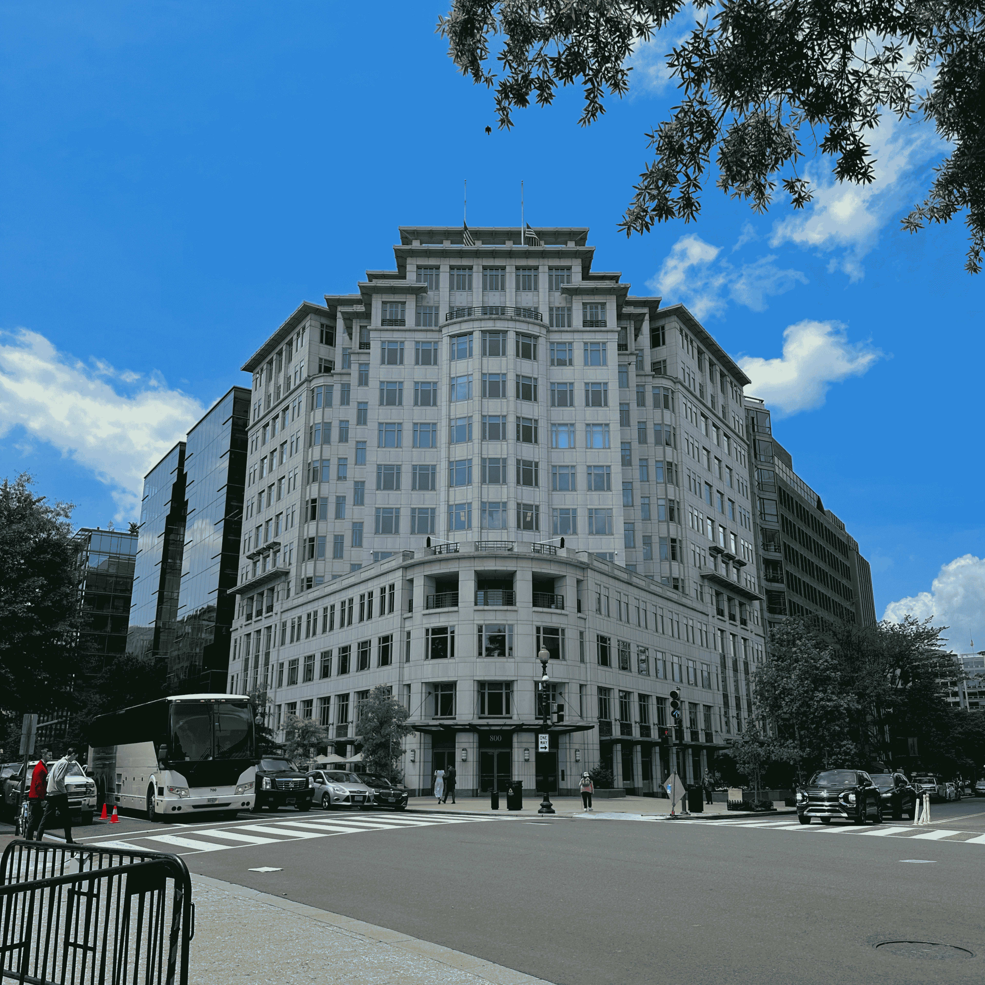 The image shows a prominent, multi-story building at an intersection with vehicles and trees in the foreground, under a clear blue sky.