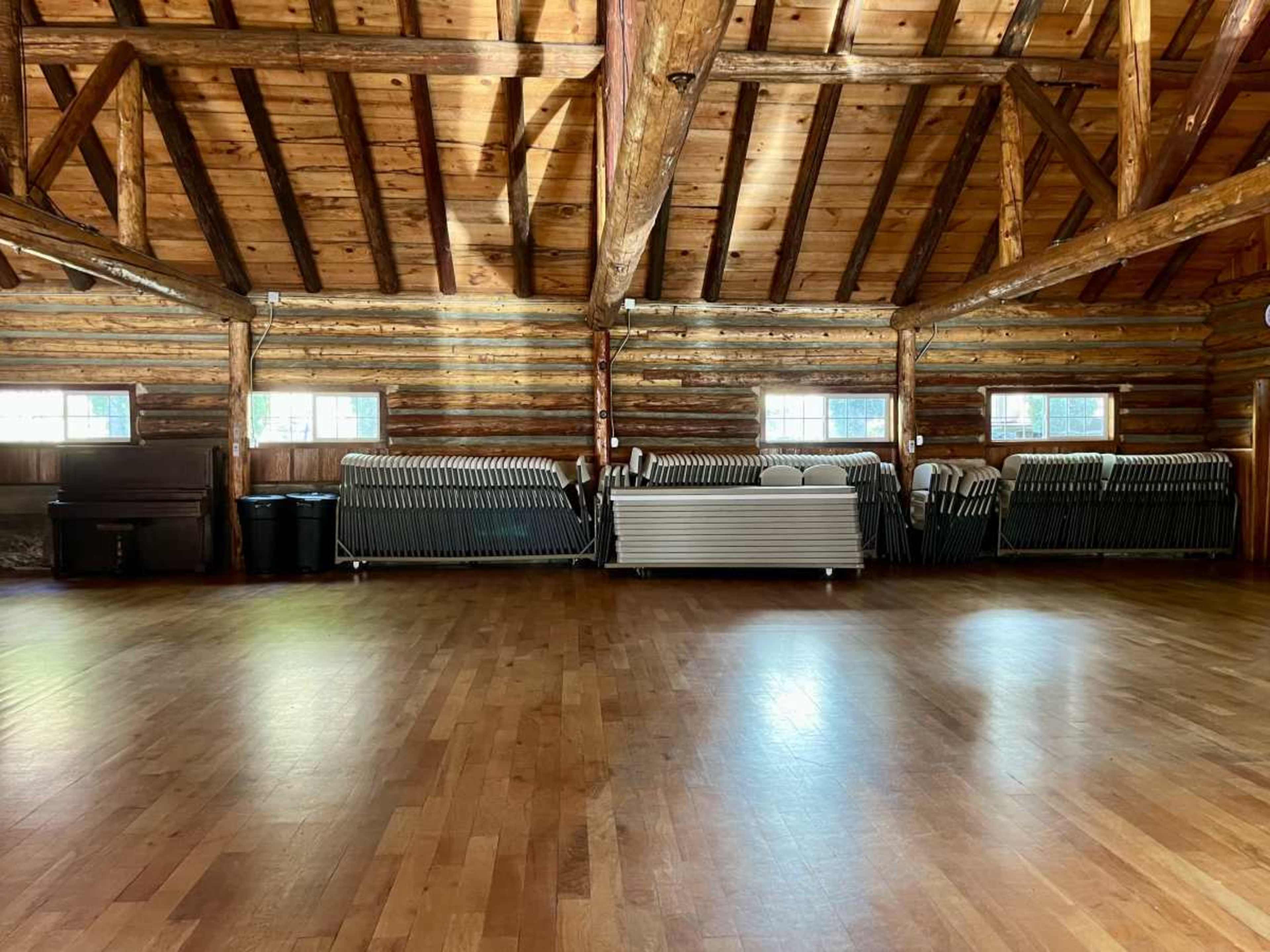 The interior of a wooden cabin features exposed beams, a polished wooden floor, and stacks of folding tables against the wall.