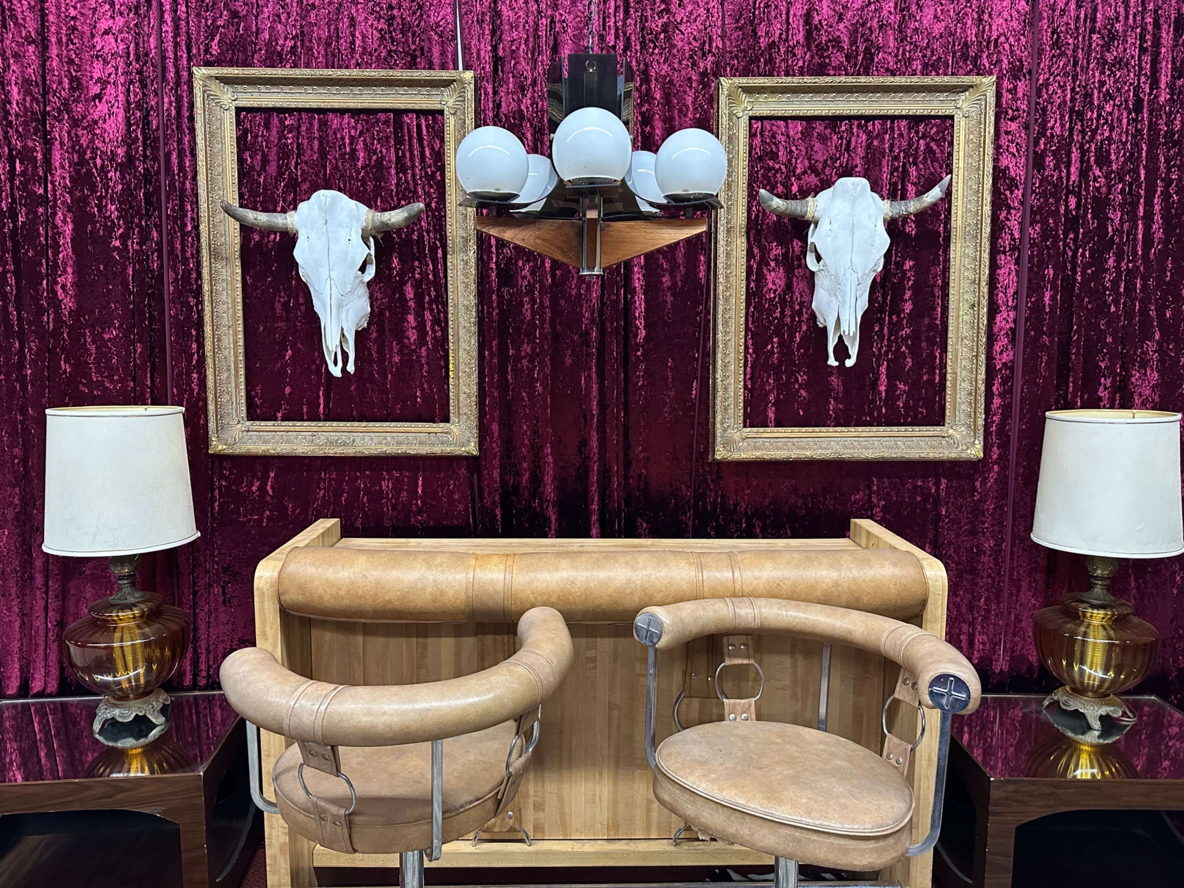 The image features a stylish bar area with two leather stools, framed bull skulls mounted on a velvet wall, and two lamps on either side.