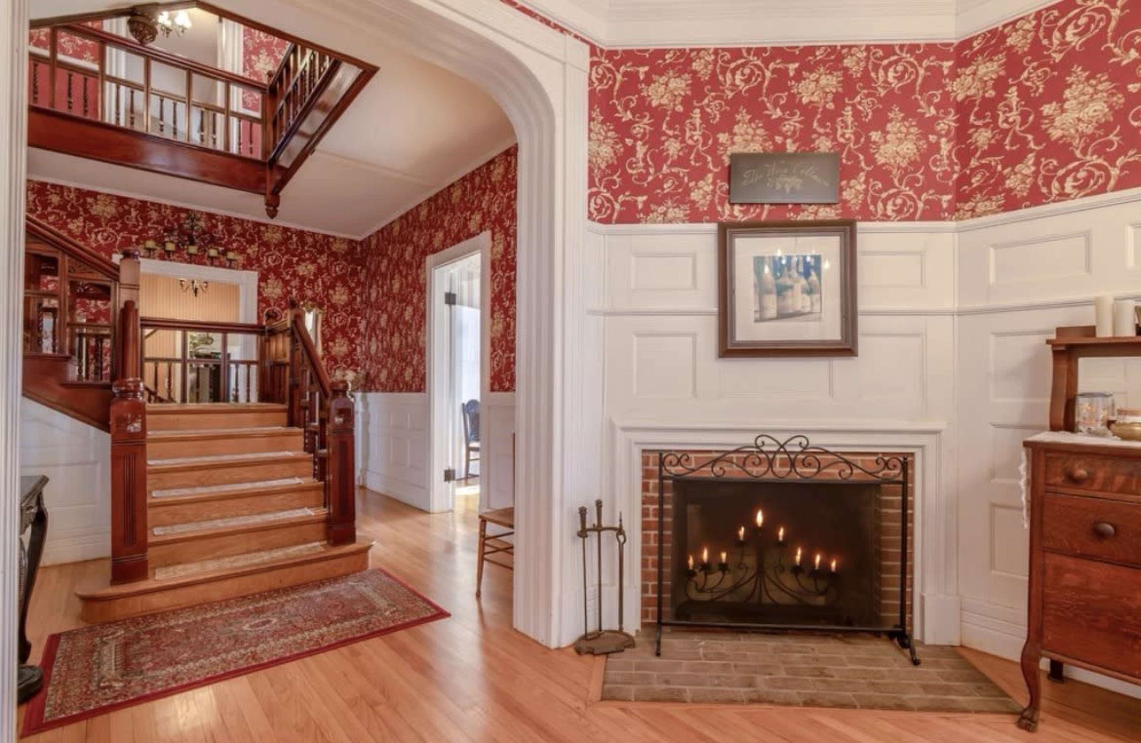 The image shows a warmly decorated entryway with red patterned wallpaper, a wooden staircase, and a fireplace in the corner.