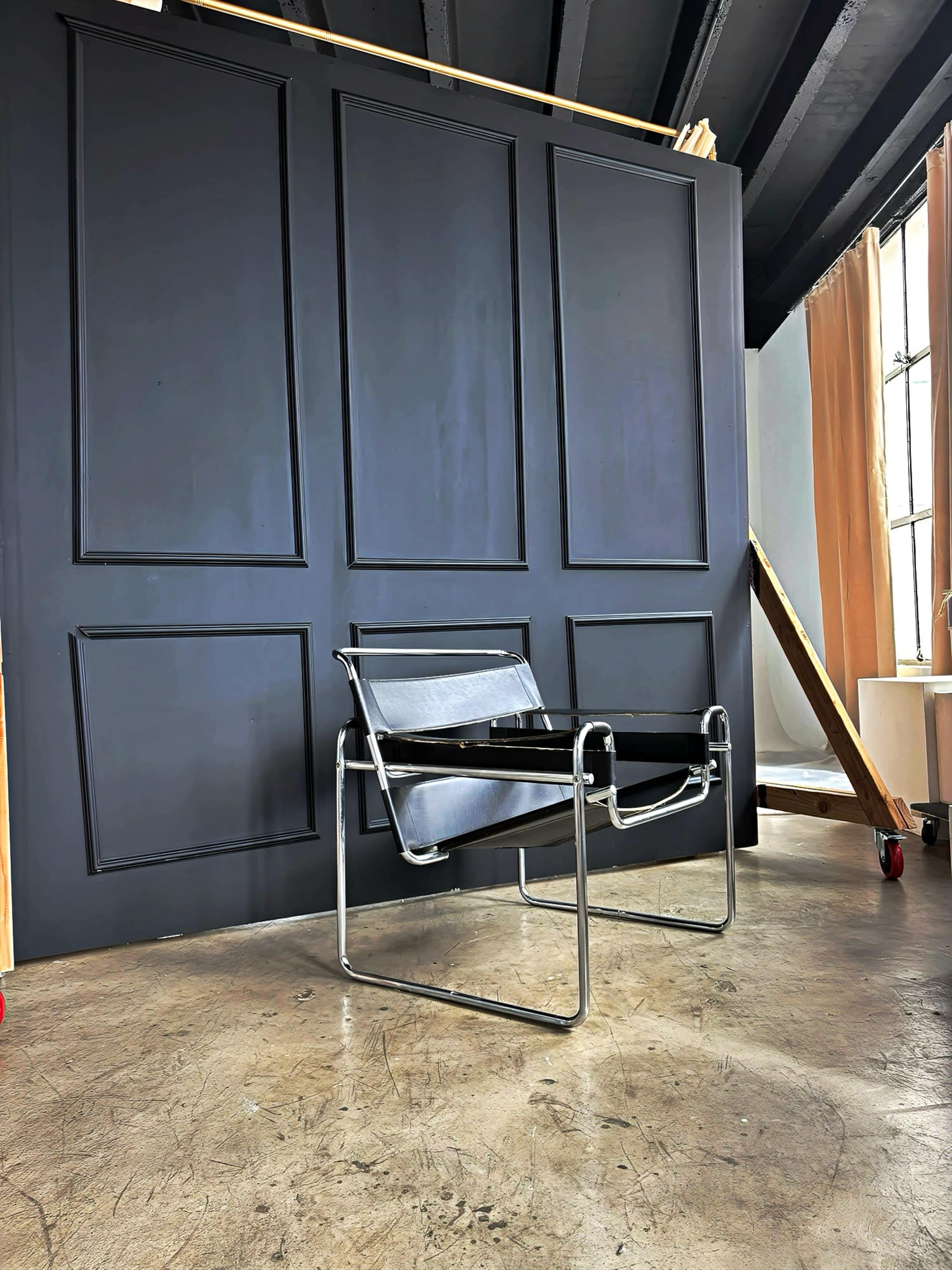 A minimalist chair with a chrome frame and black seat is positioned in front of a dark paneled wall in an interior space.