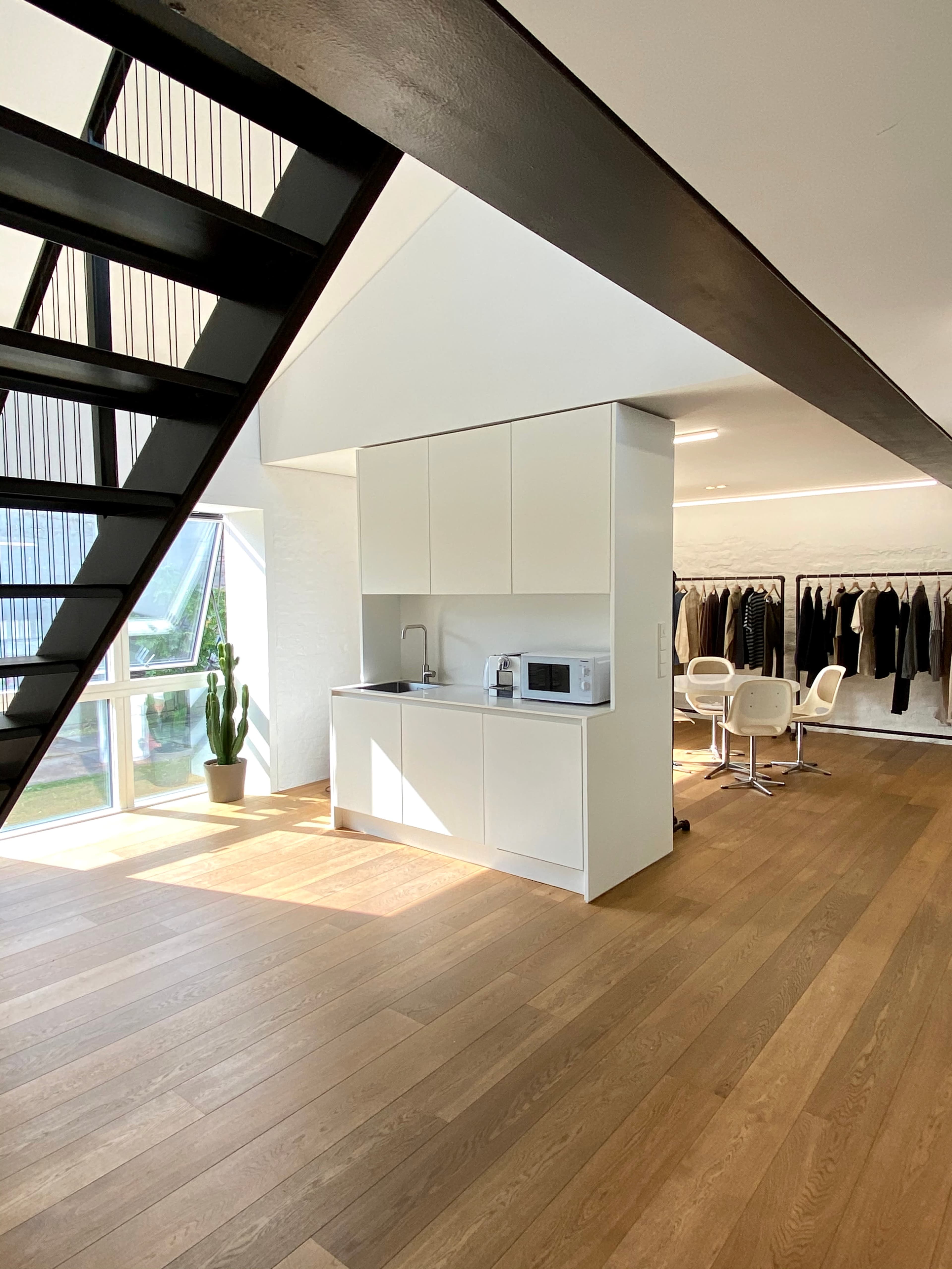 A modern kitchen with white cabinetry and a microwave, alongside a clothing rack with various garments in a well-lit room featuring wooden flooring.