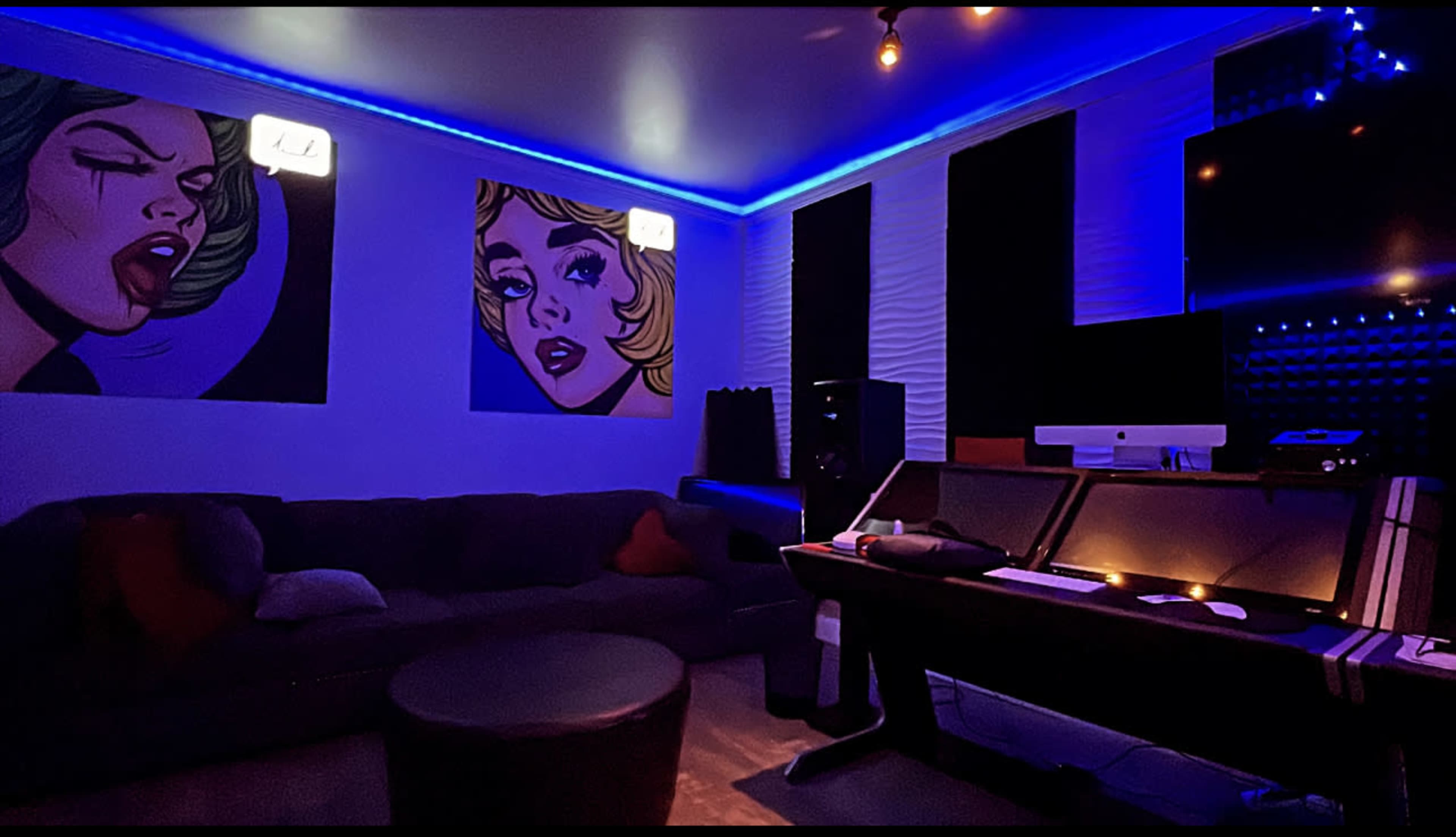 A modern music studio with colorful wall art, a dark couch, and a desk equipped with audio equipment under blue LED lighting.