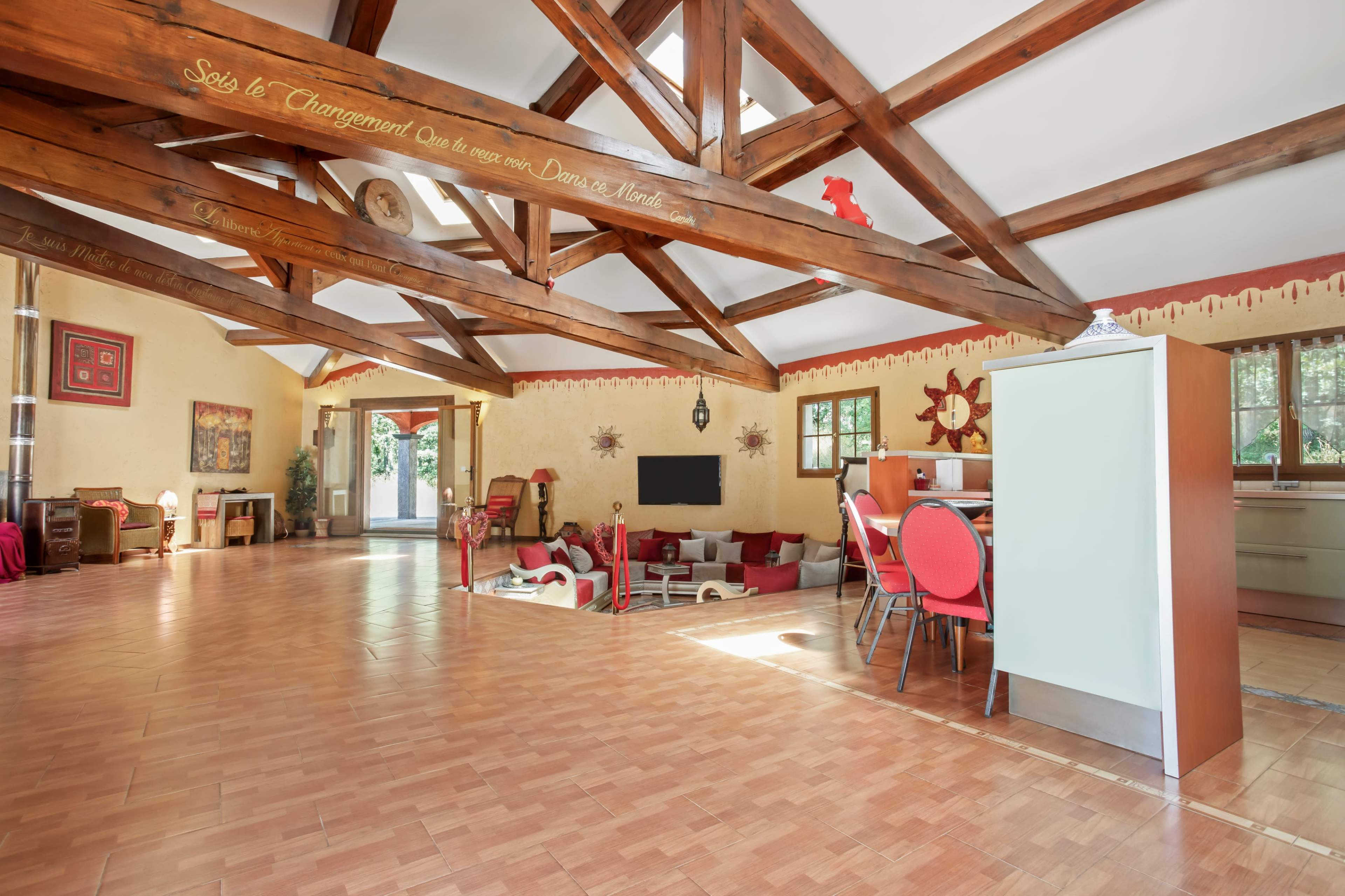 The image shows a spacious interior room with exposed wooden beams, a large open area, and a combination of seating and kitchen spaces.
