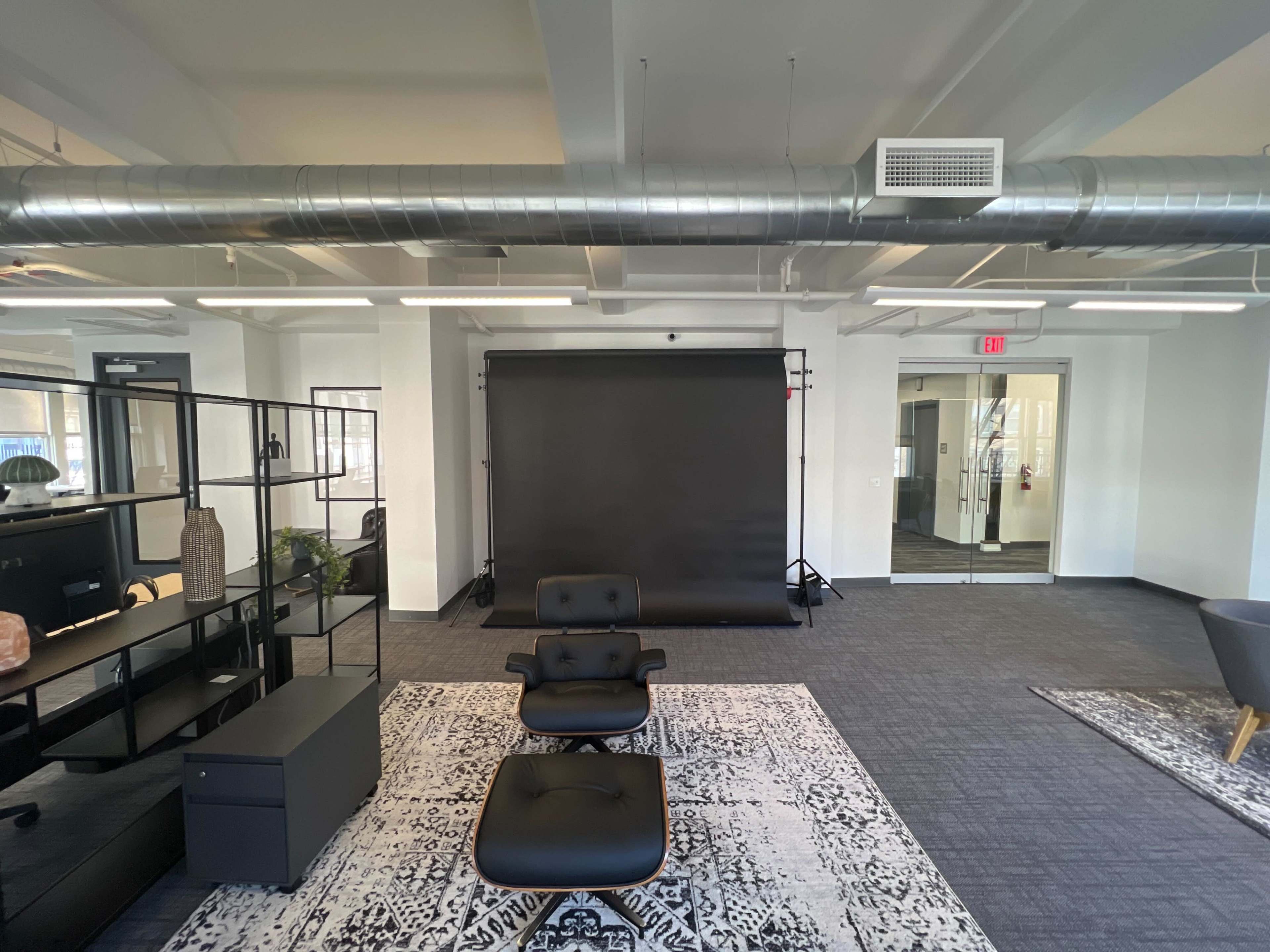 A modern office space featuring a black lounge chair, a patterned rug, and a backdrop setup against a white wall.