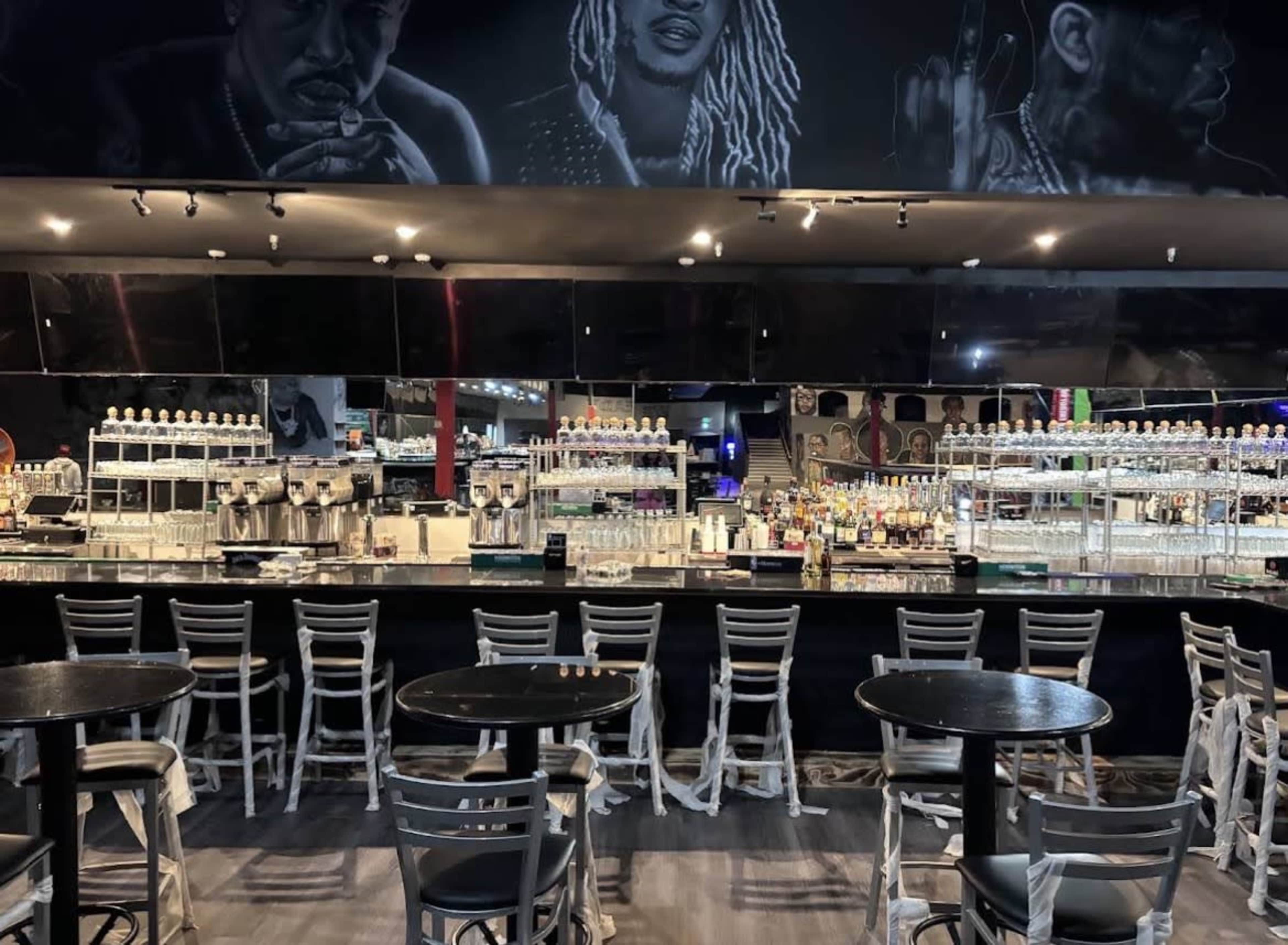 The image shows a bar area with multiple bottles displayed behind the counter and empty tables arranged in front.