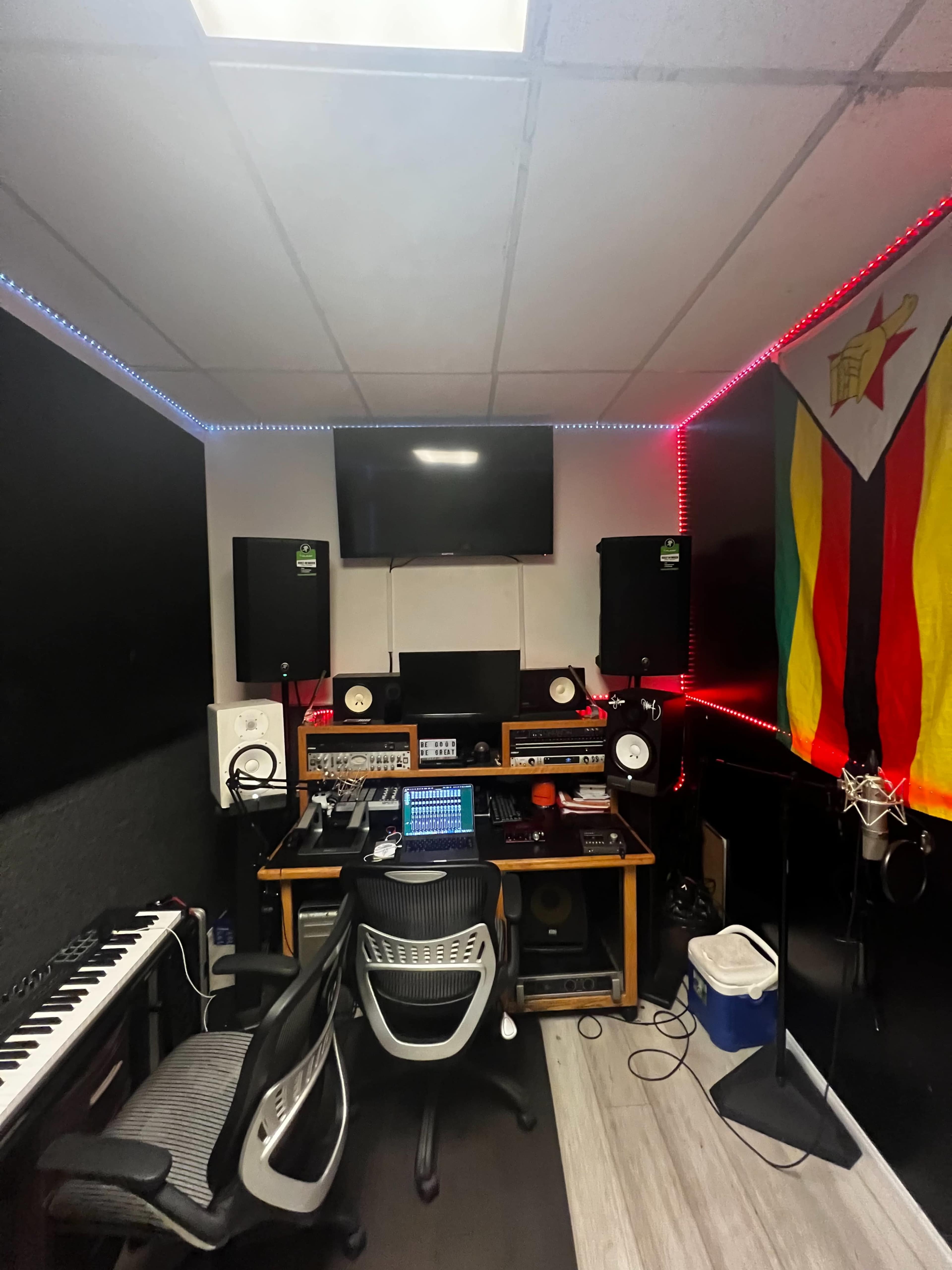 A music recording studio featuring a desk with audio equipment, speakers, a computer, and a microphone, along with a Zimbabwean flag on the wall.