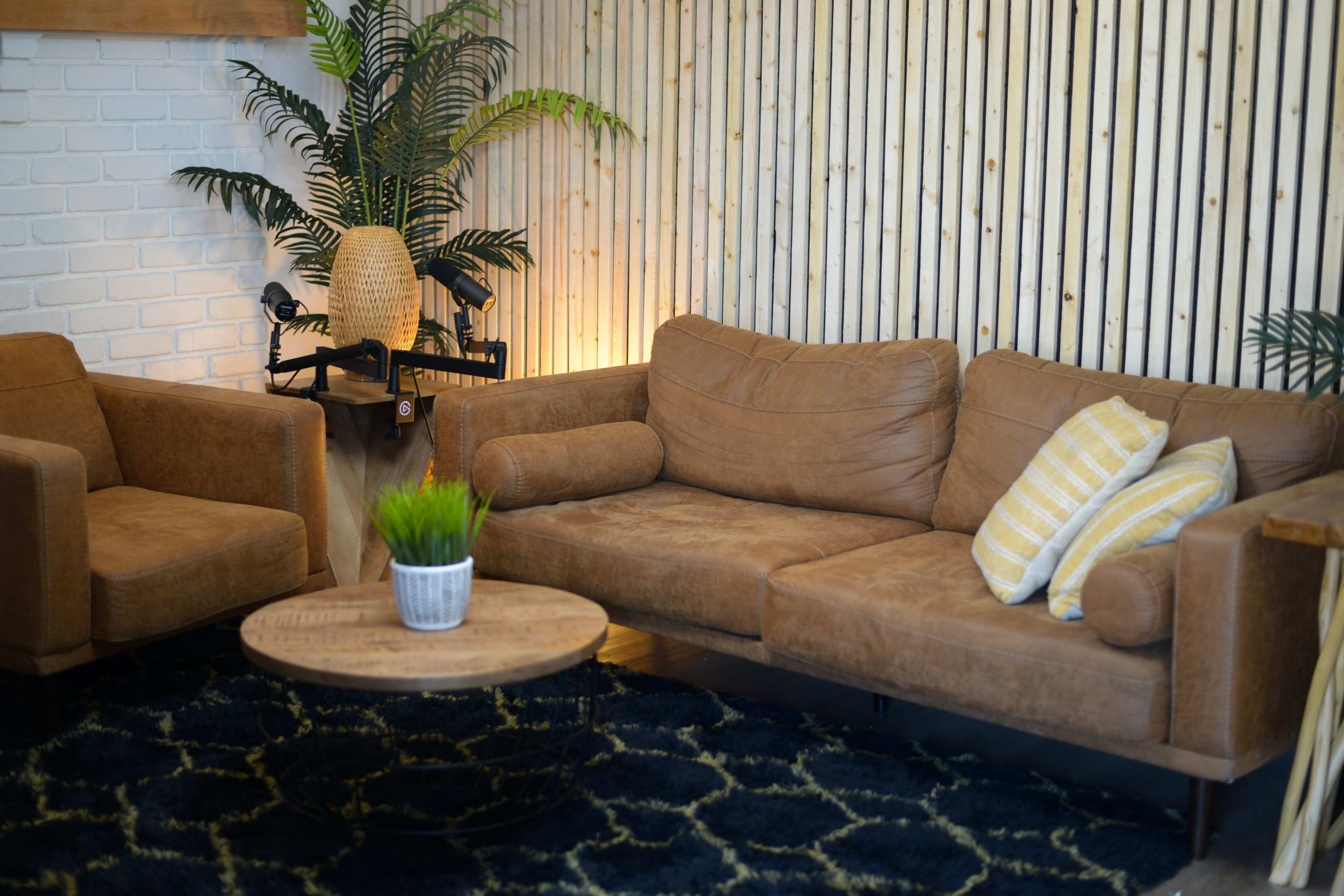 A cozy living area with a brown leather sofa and armchair, a circular wooden coffee table, and decorative plants against a striped wooden wall.