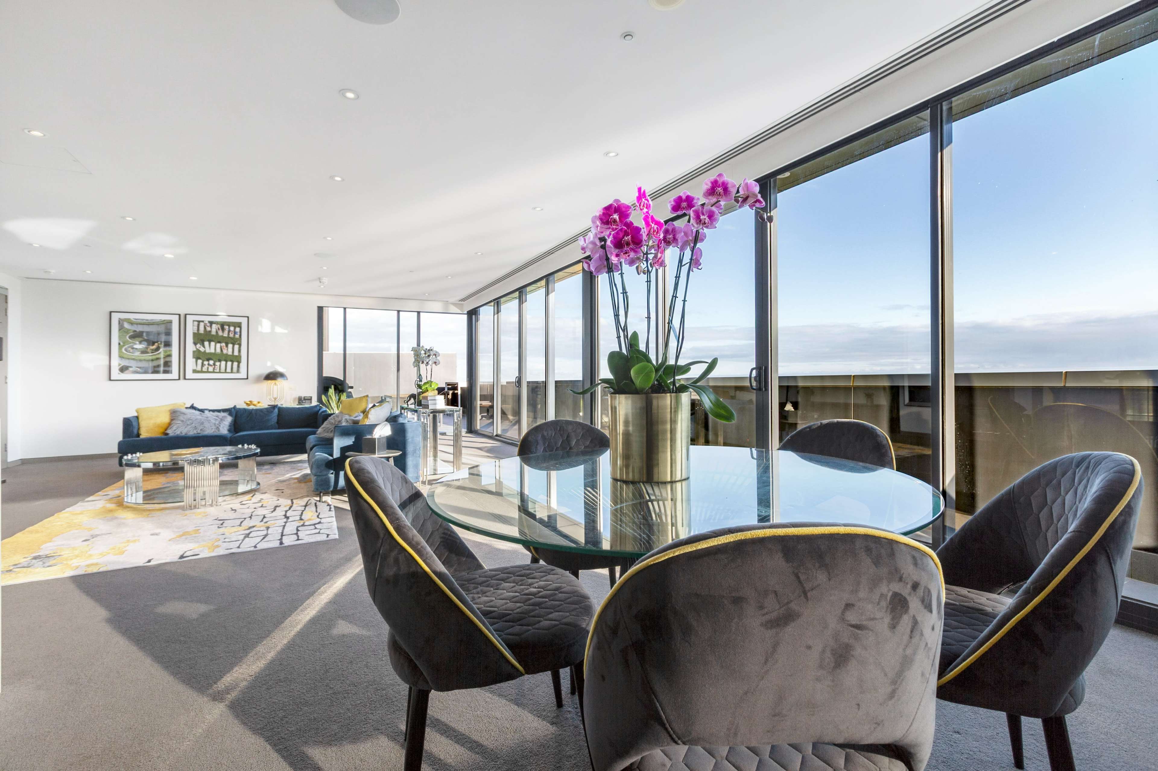 The image shows a modern living space featuring a dining table with four chairs, a sectional sofa, and large windows providing panoramic views.