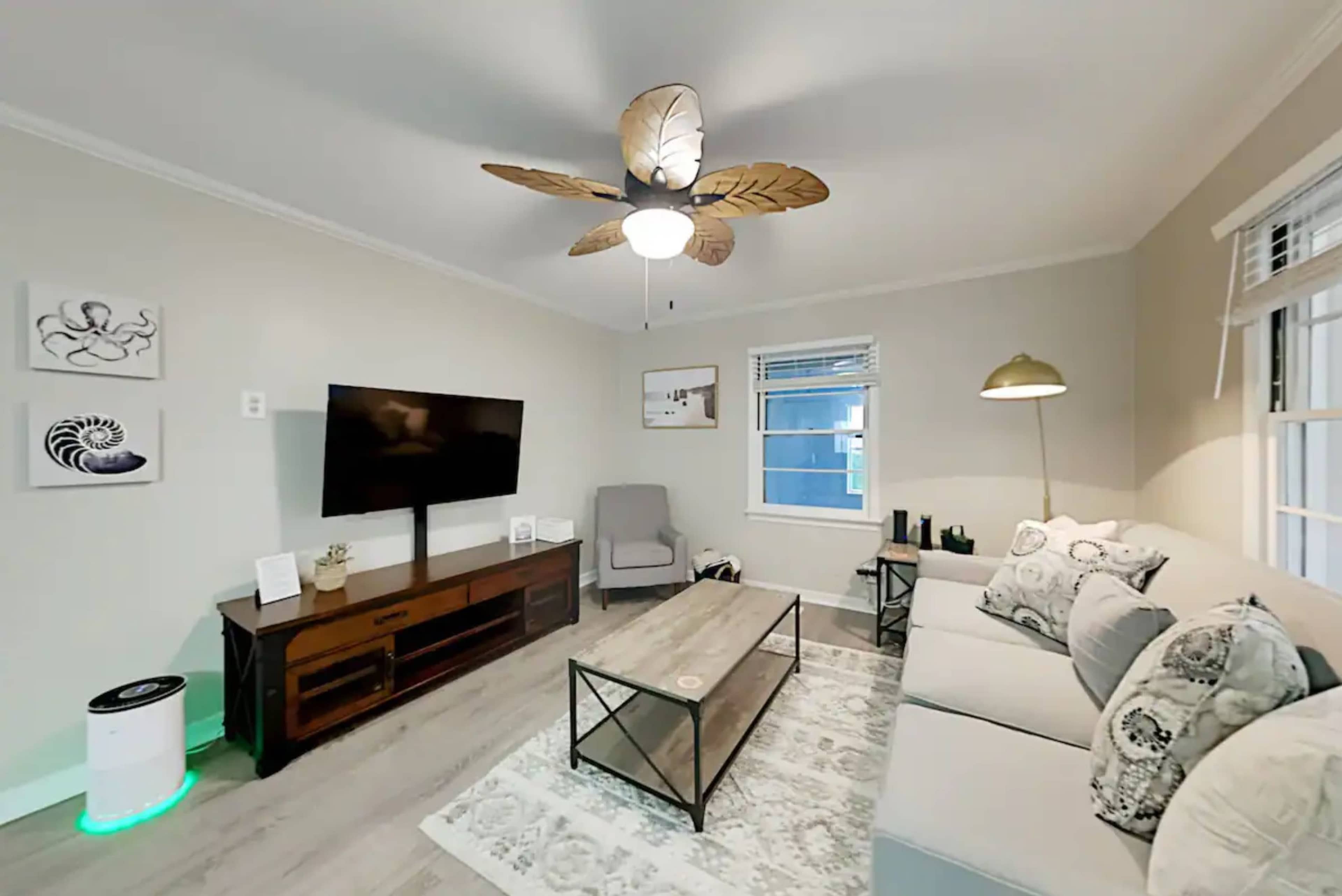 The living room features a sectional sofa, a coffee table, a television mounted on the wall, and a ceiling fan with leaf-shaped blades.