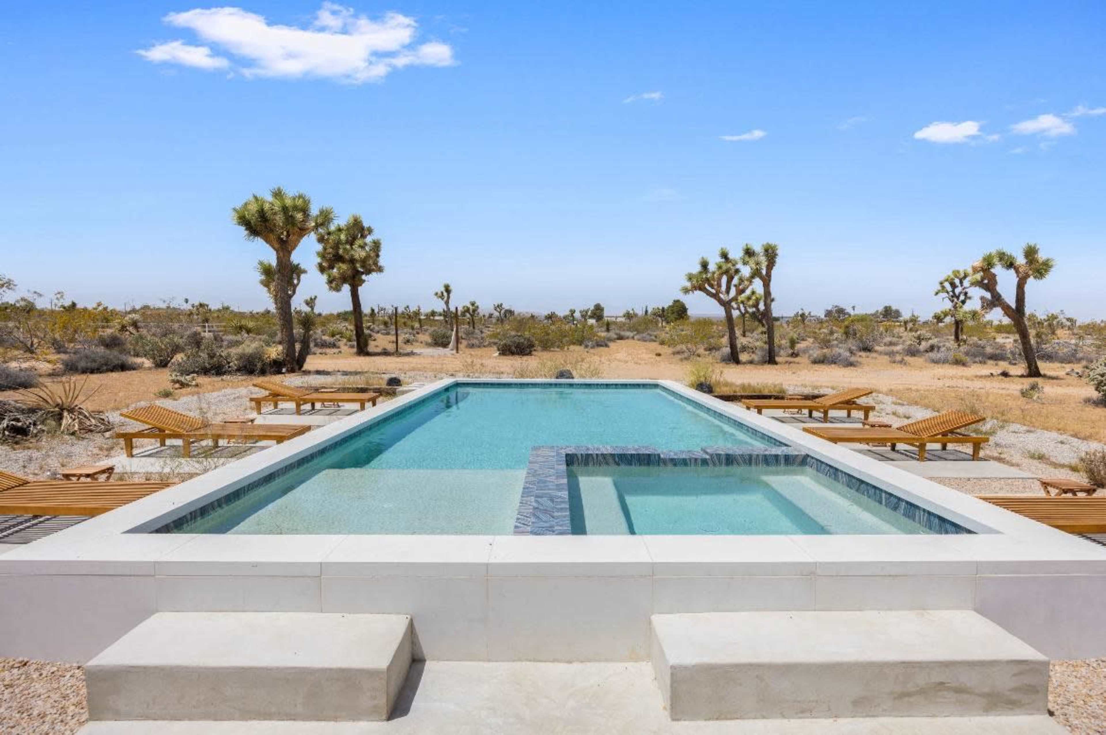 A rectangular swimming pool with a spa, surrounded by desert landscape and Joshua trees.