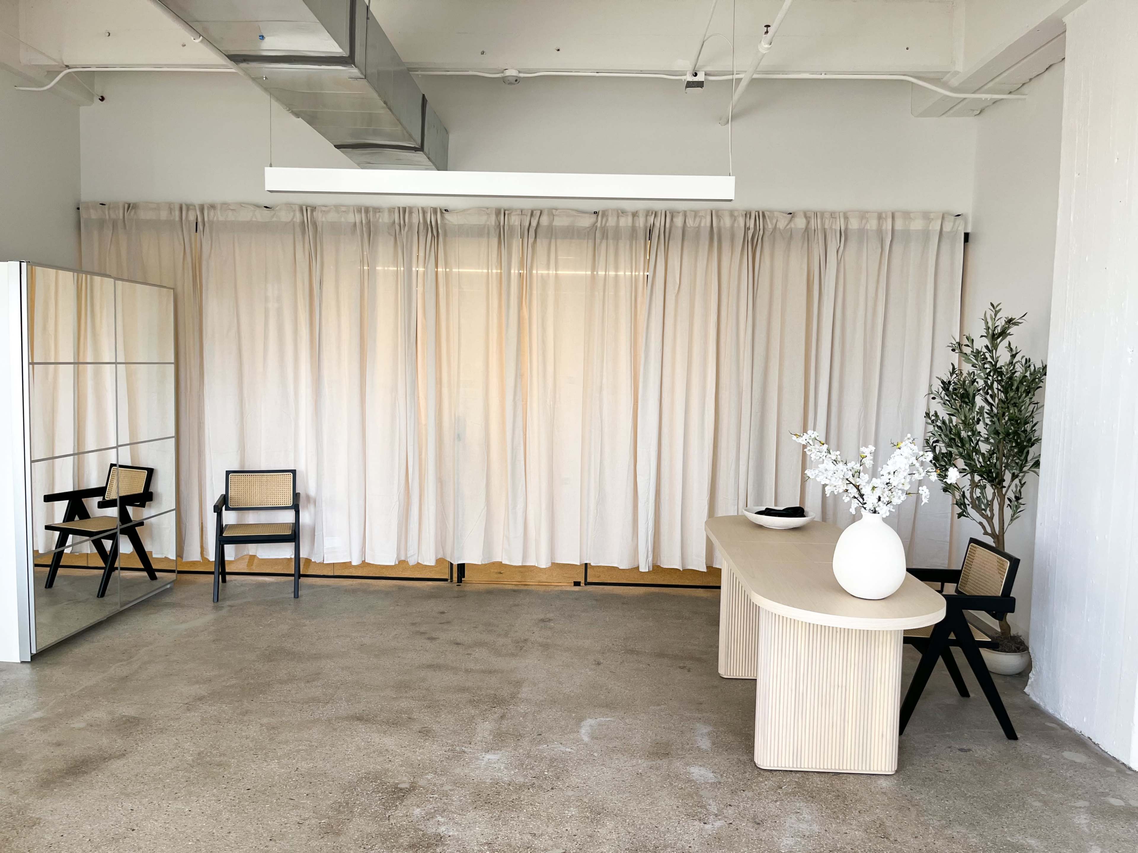 A minimalistic interior with a wooden table, two black chairs, a large mirror, and sheer curtains, creating an open and airy space.