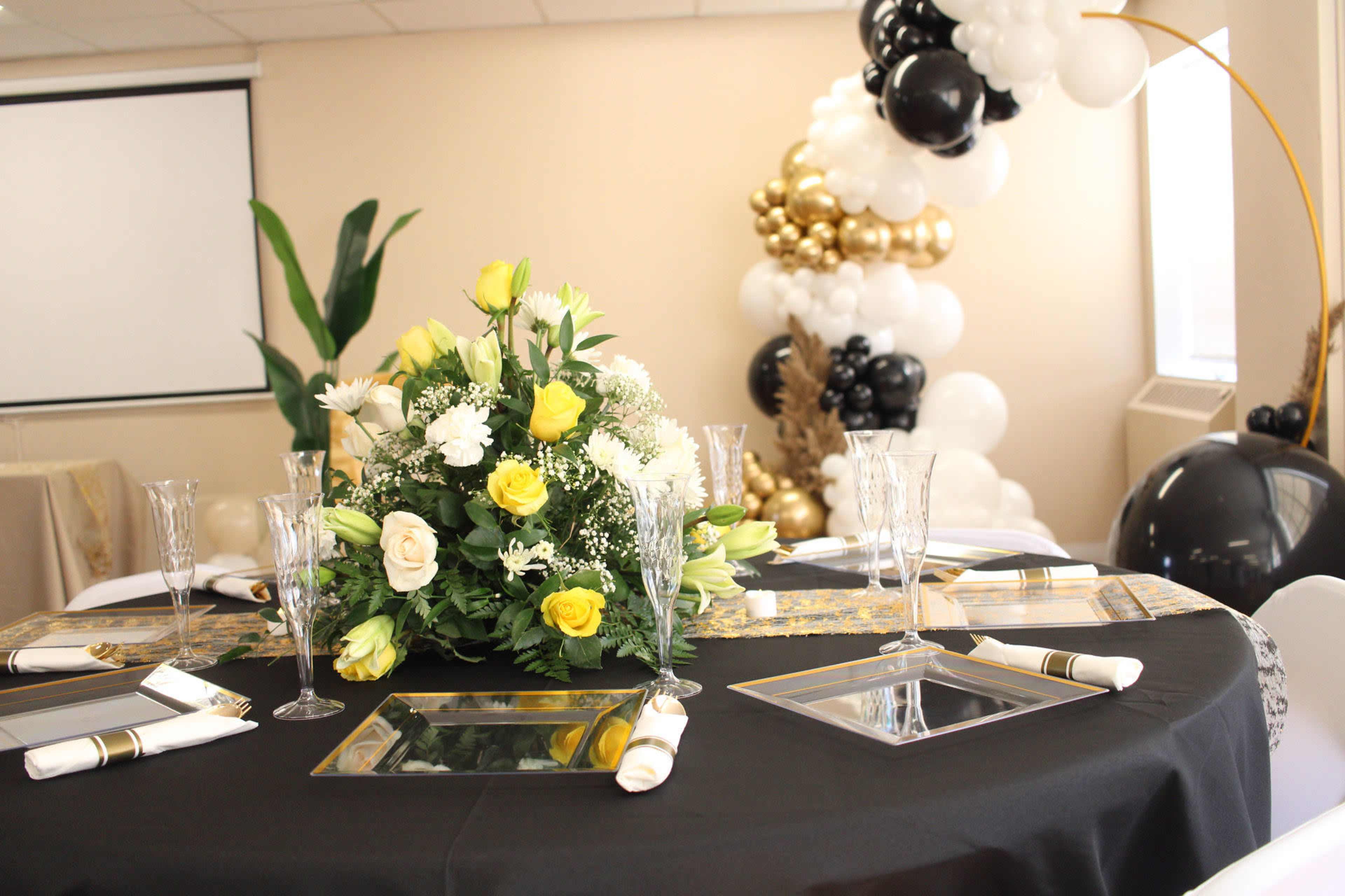 A circular dining table is set with a floral centerpiece of yellow and white flowers, surrounded by elegant glassware and a black tablecloth, with a backdrop of decorative balloons in gold, white, and black.