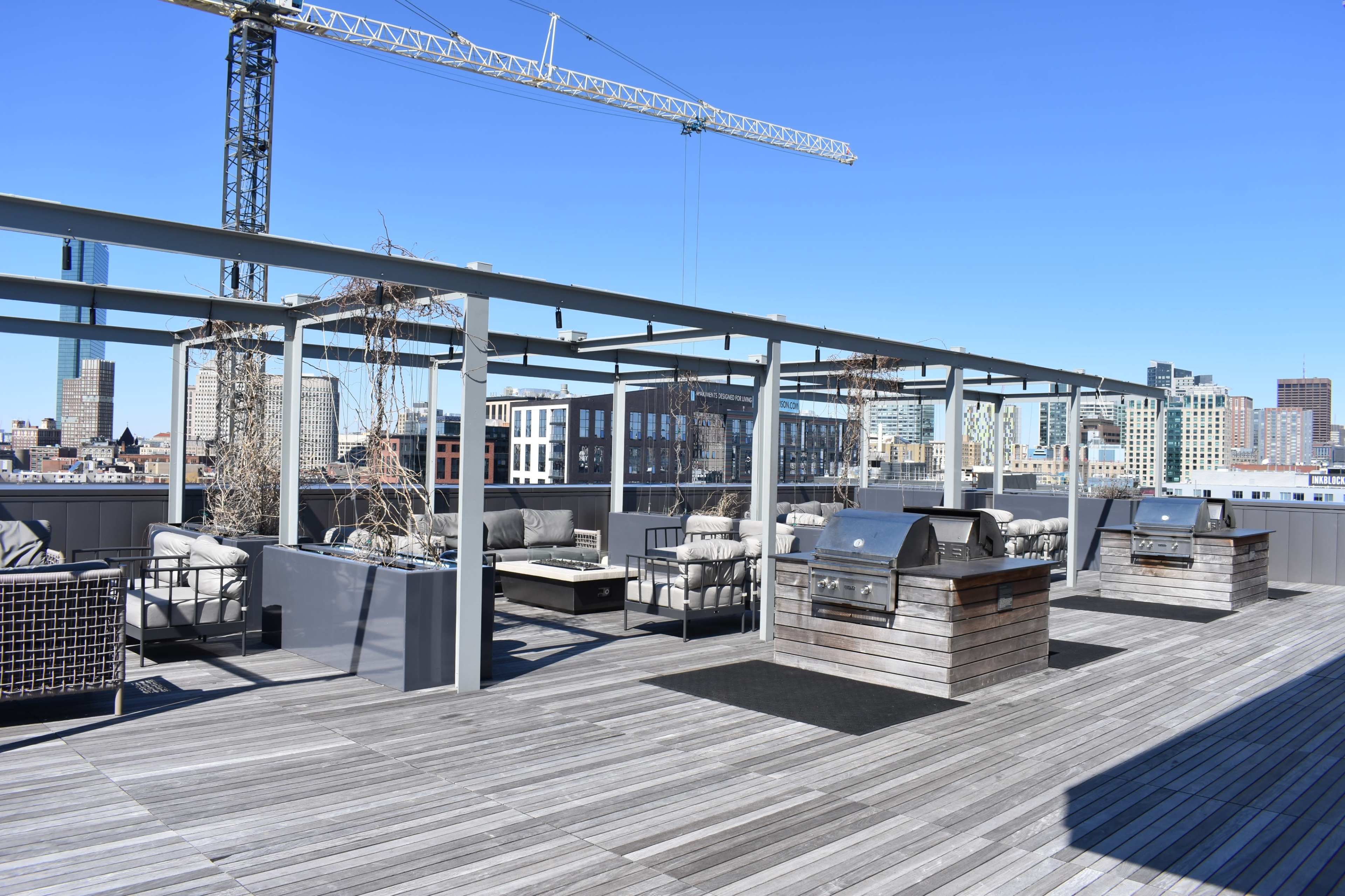 A rooftop deck featuring outdoor seating arrangements, barbecue grills, and a skyline view of a city under a clear blue sky.