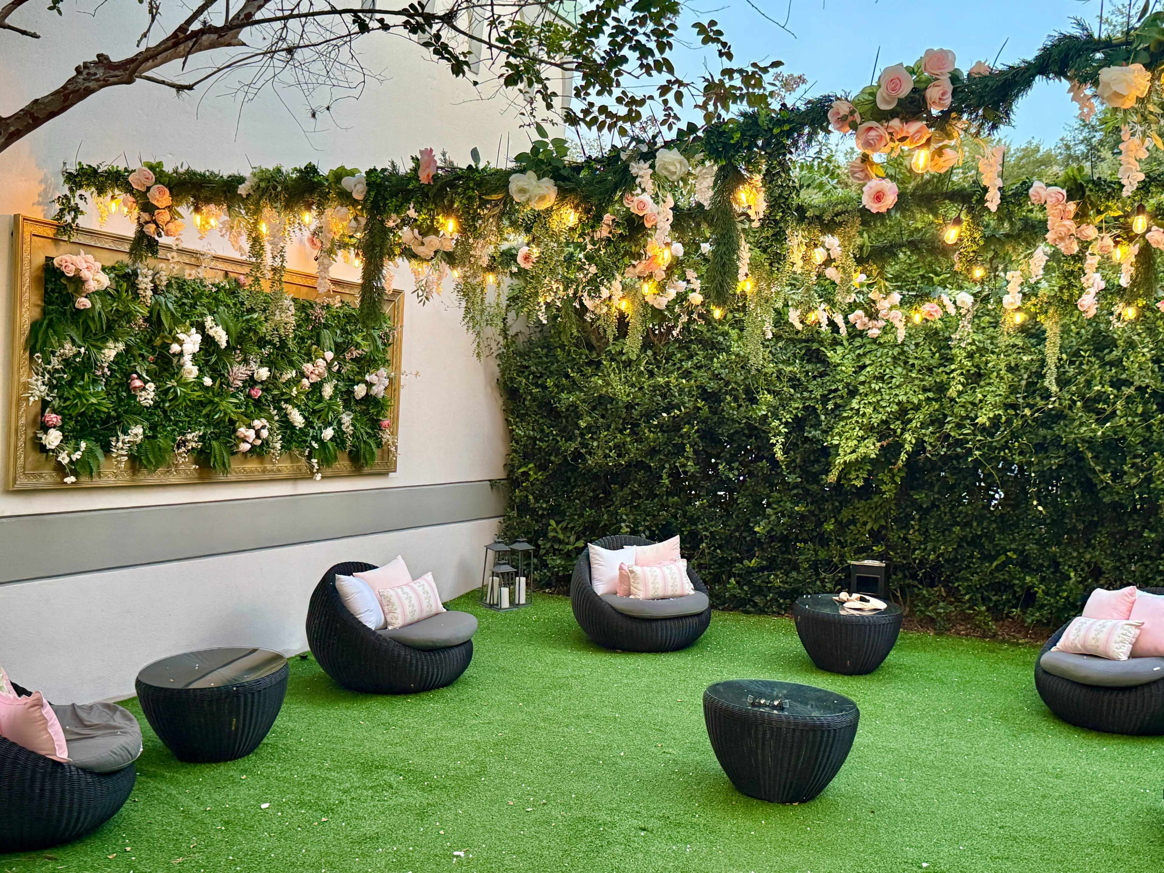 The scene features a cozy outdoor lounge area with round black chairs, small tables, and a backdrop of greenery and floral decorations illuminated by string lights.