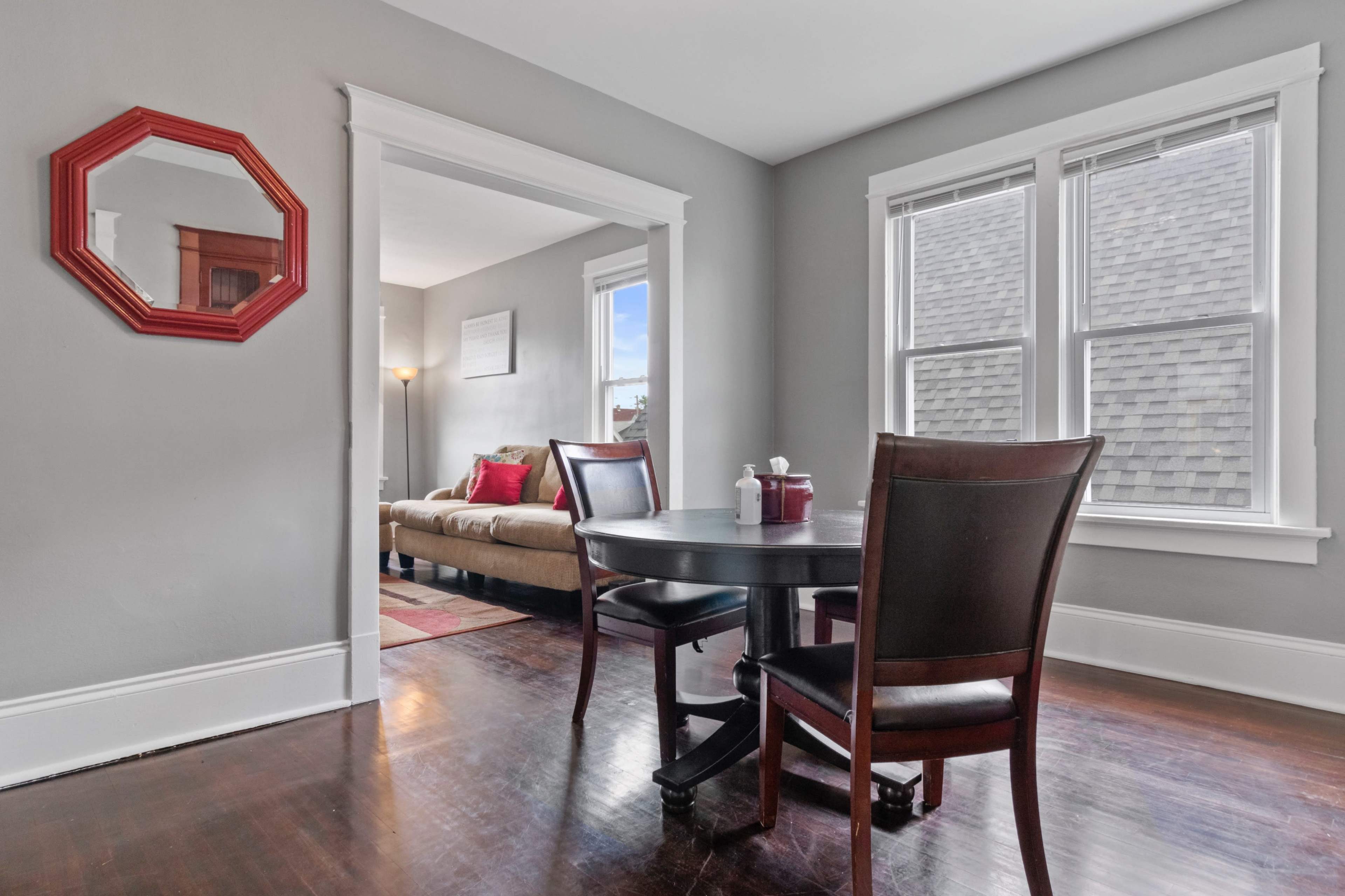 A dining area with a round black table and two chairs is visible, framed by a doorway that leads to a bright living room with large windows.