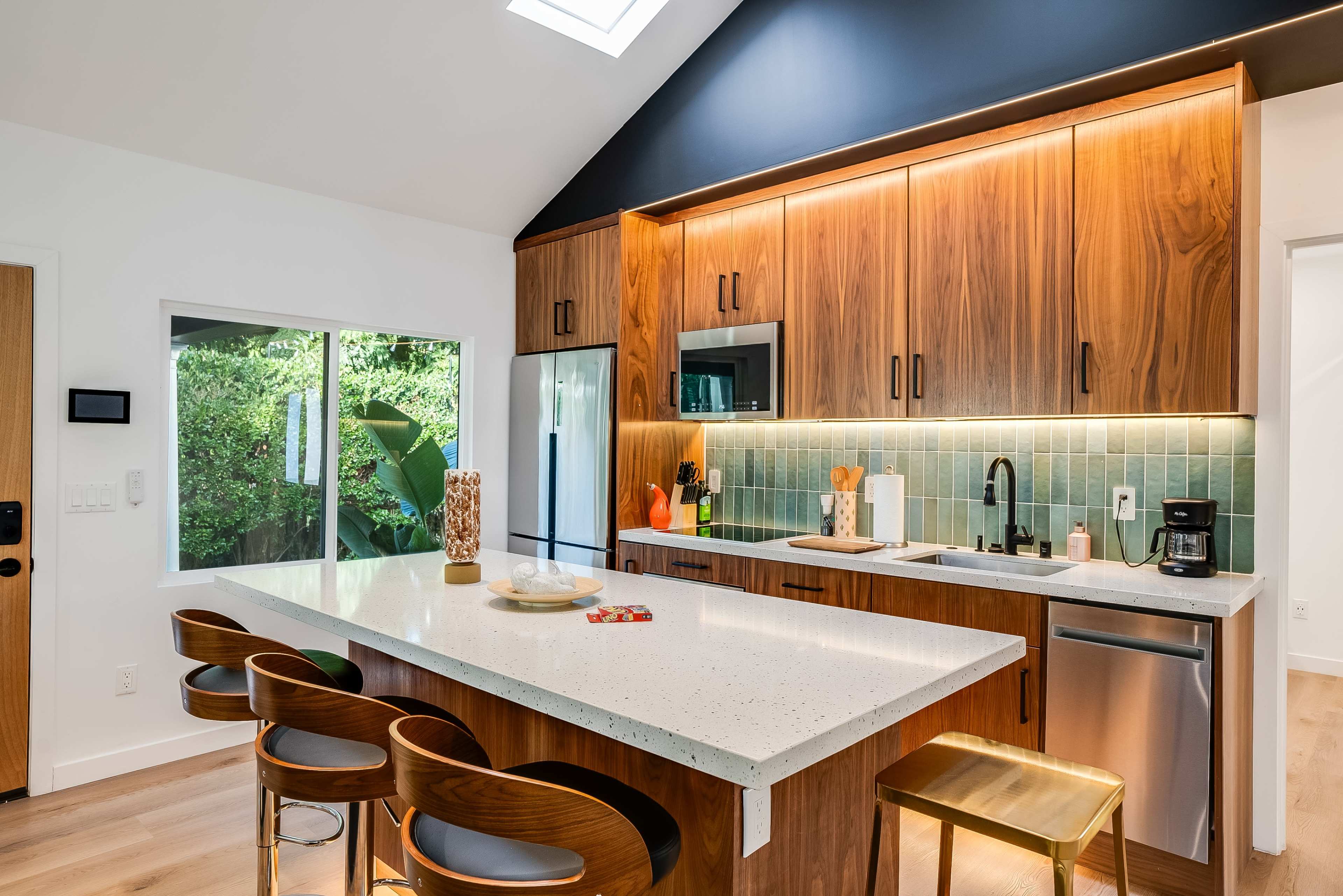 A modern kitchen features wooden cabinets, a marble island with barstools, and a large window providing views of greenery outside.