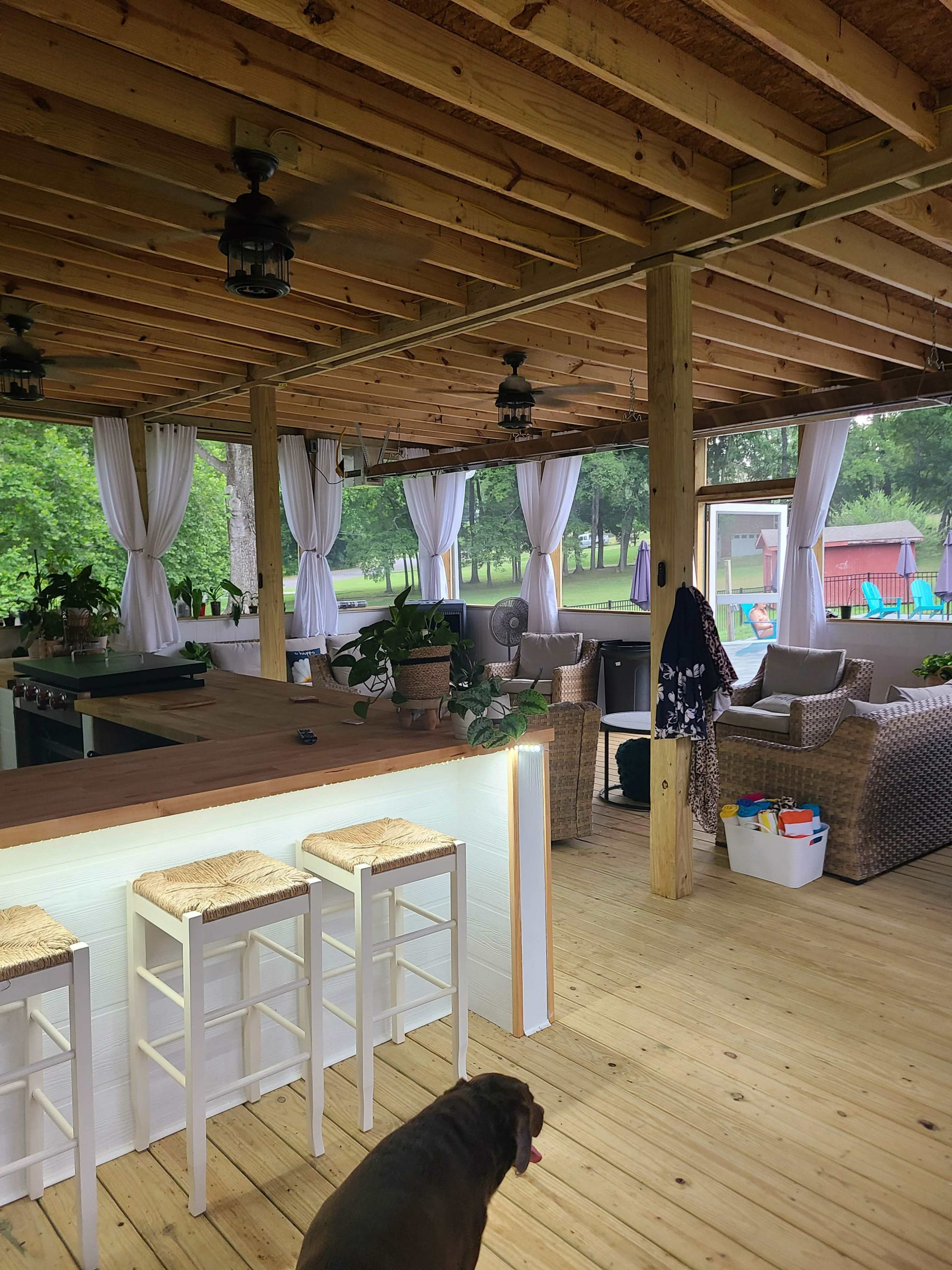 The image shows a spacious outdoor patio area with wooden beams, comfortable seating, and decorative plants, along with a bar counter and visible green surroundings.