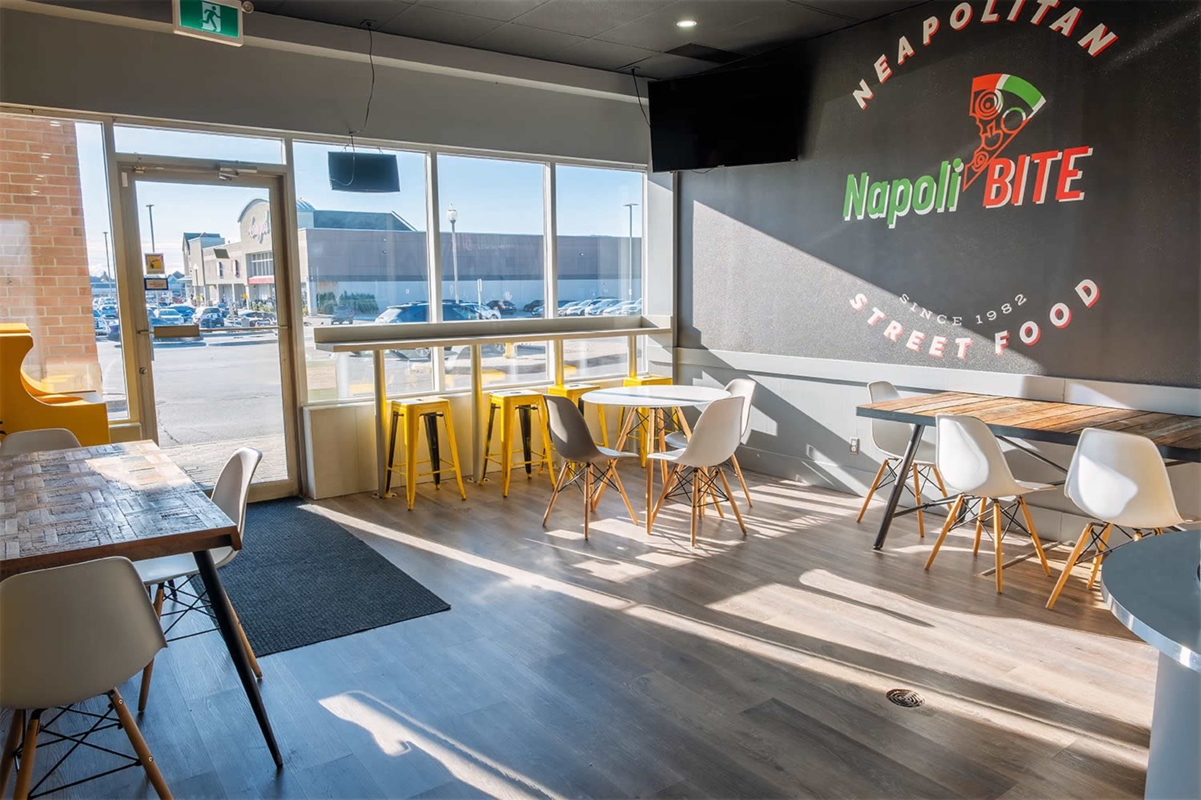 A modern café interior featuring wooden tables, white chairs, large windows, and a vibrant wall sign for Napoli Bite Street Food.