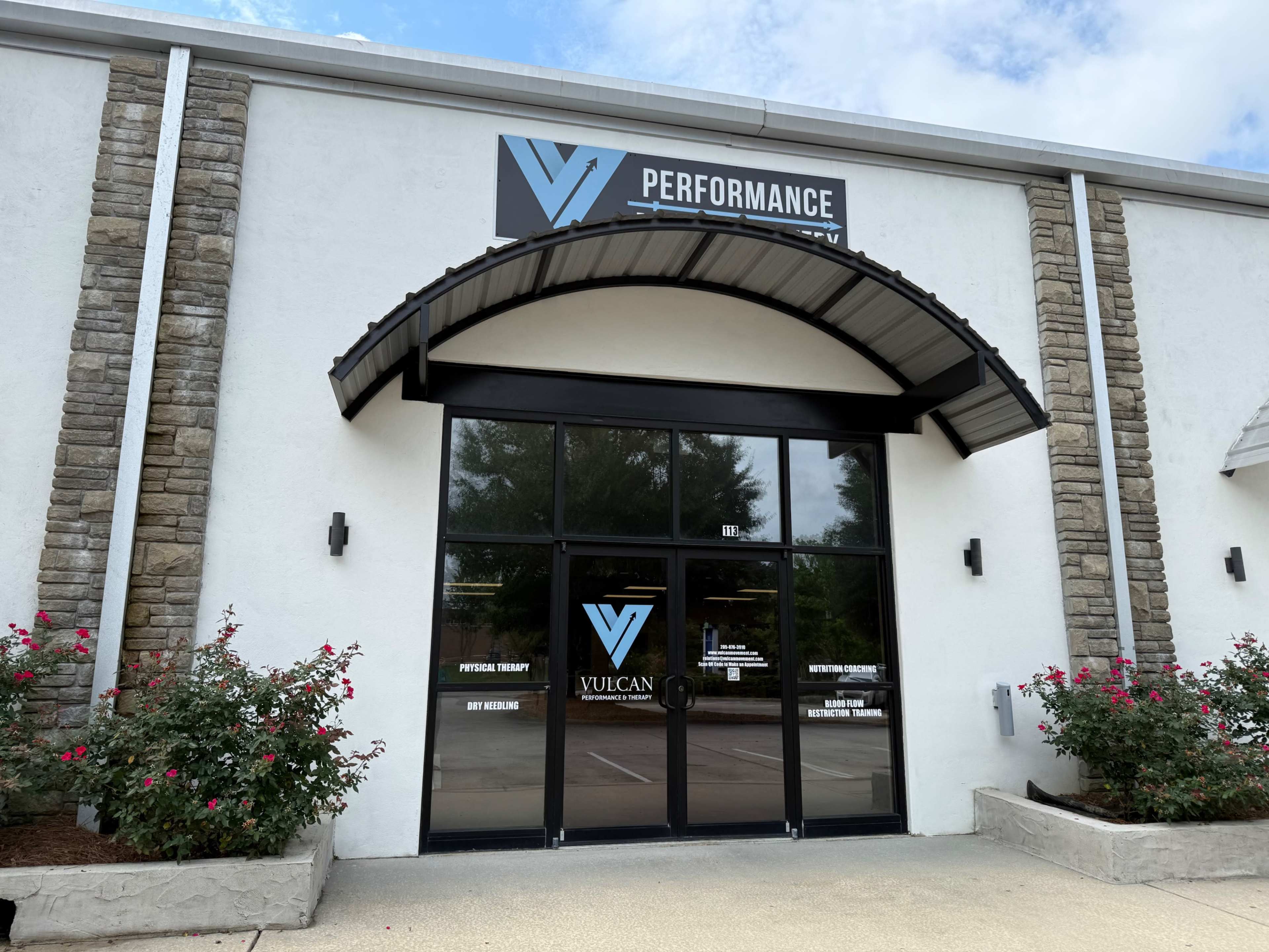 The image shows a commercial building entrance with a sign that reads "VULCAN PERFORMANCE" above the glass doors, framed by stone accents and surrounded by rose bushes.