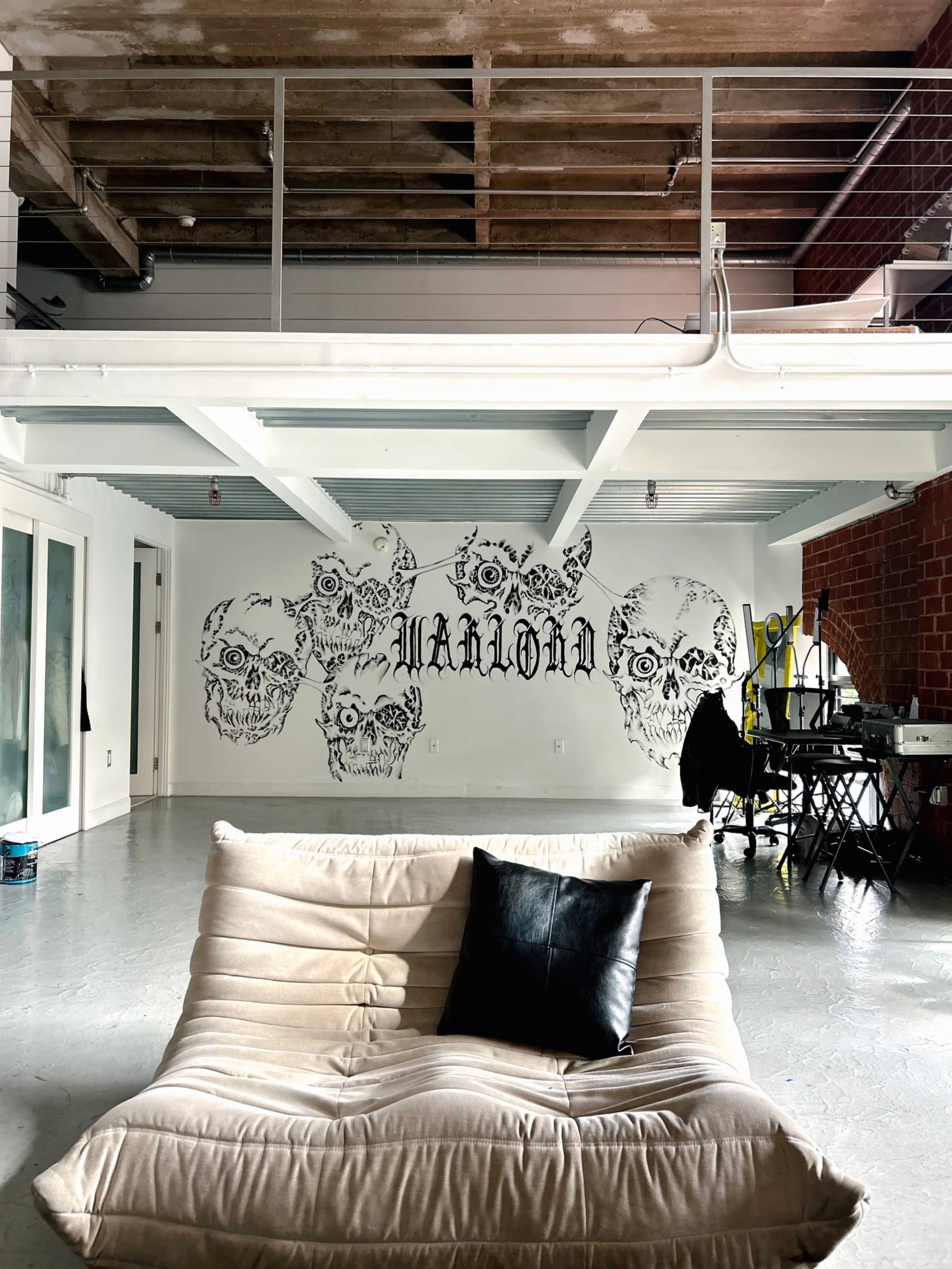 The image shows a spacious, industrial-style room with exposed wooden beams and a large mural of skulls on the wall, featuring a soft couch in the foreground.