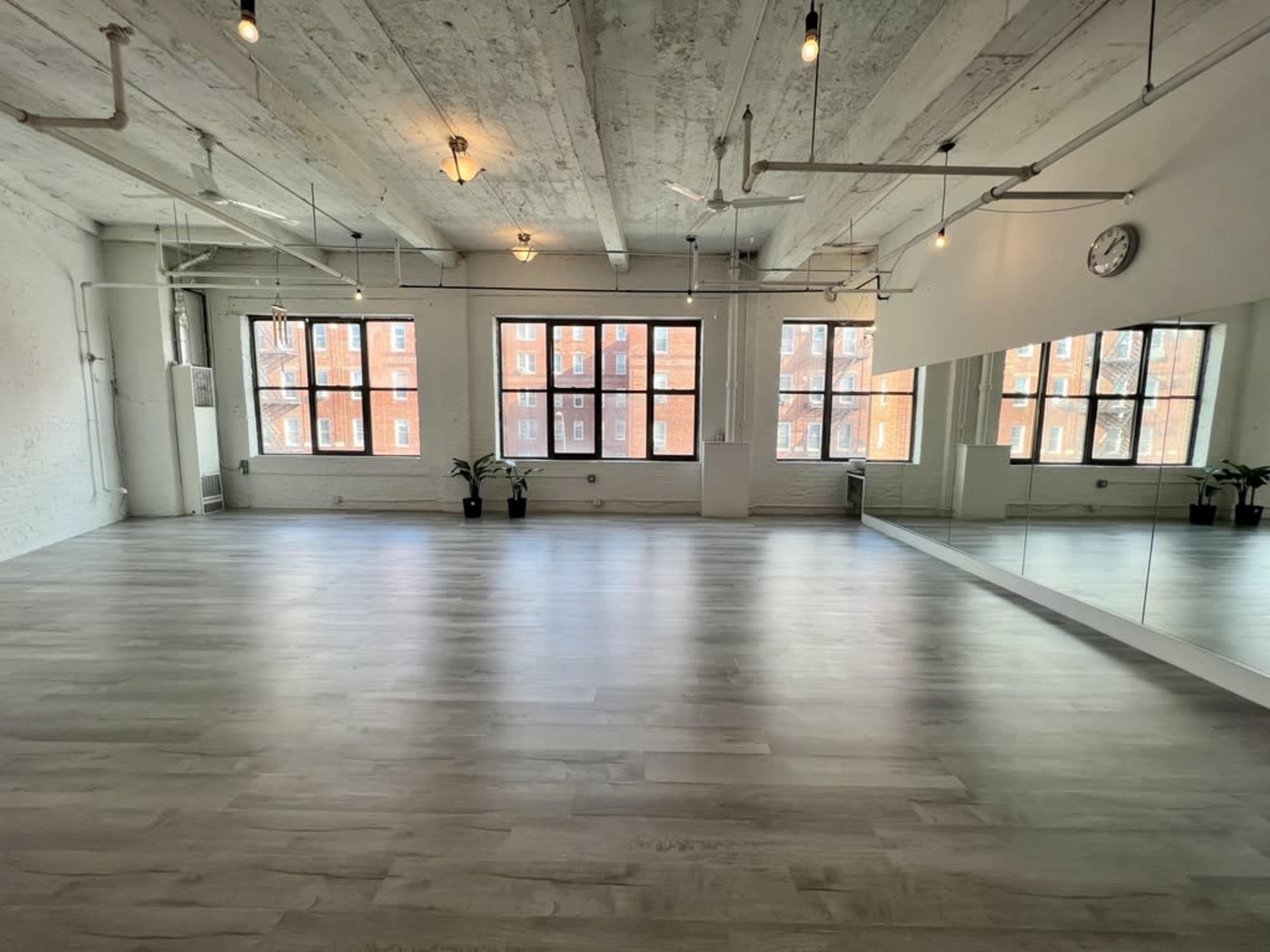 The image shows a spacious, well-lit room with large windows, wooden flooring, and a mirrored wall.