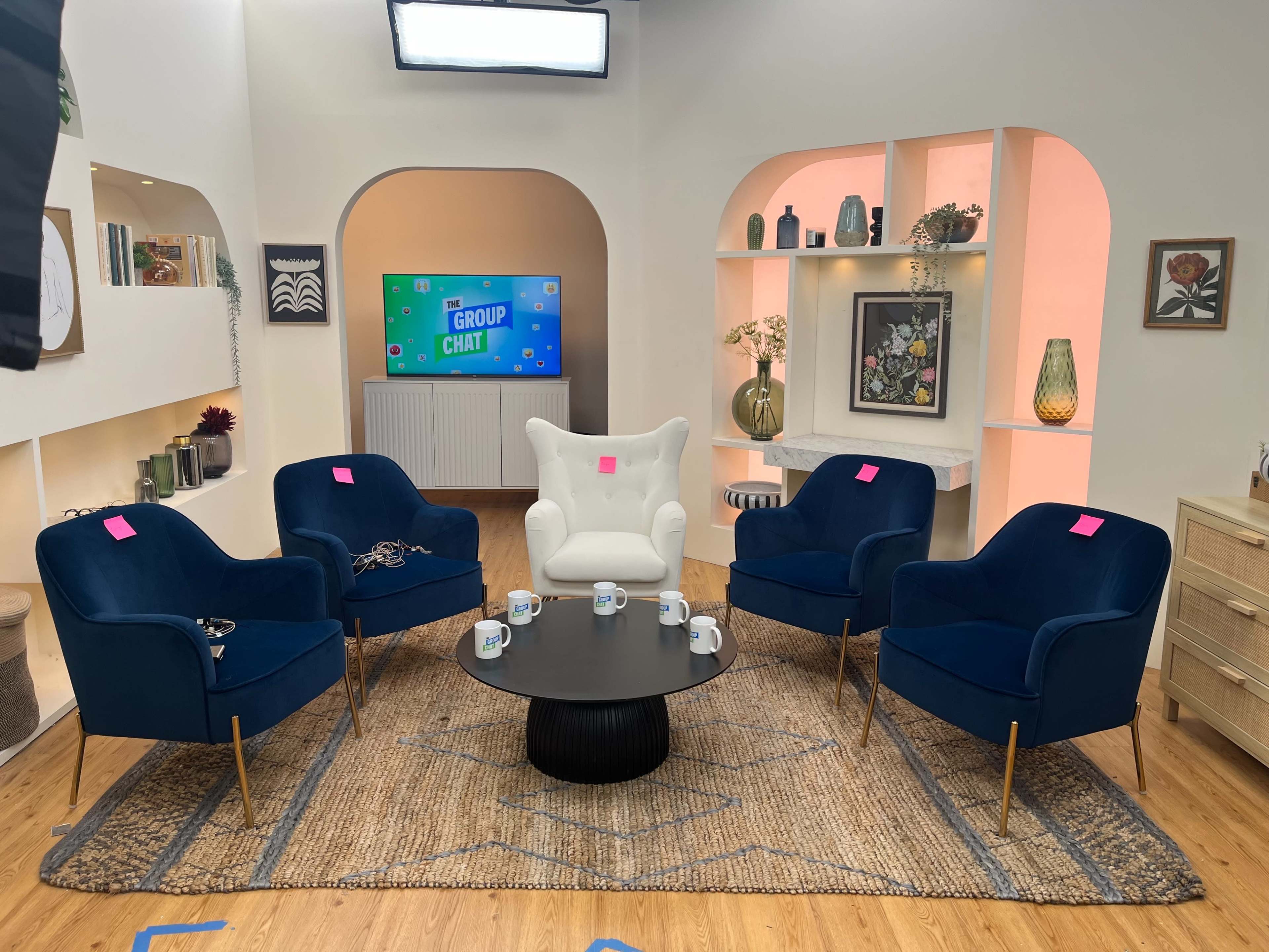 The image features a modern talk show set with five chairs arranged around a circular table on a wooden floor, surrounded by decorative shelves and a television screen displaying "GROUP CHAT."