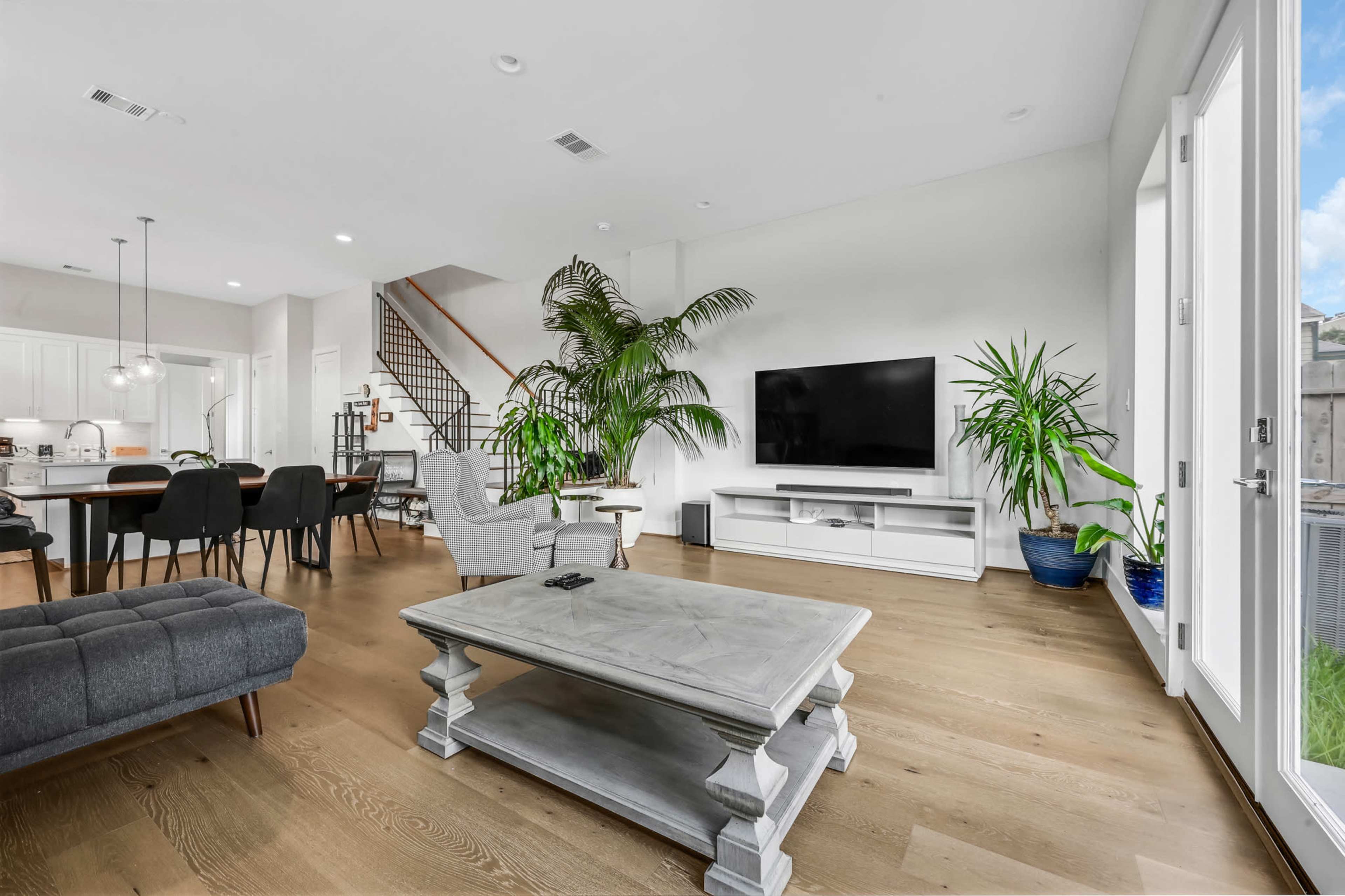 A modern living room features a large television on a white media console, a grey couch, and potted plants, with a staircase visible in the background.