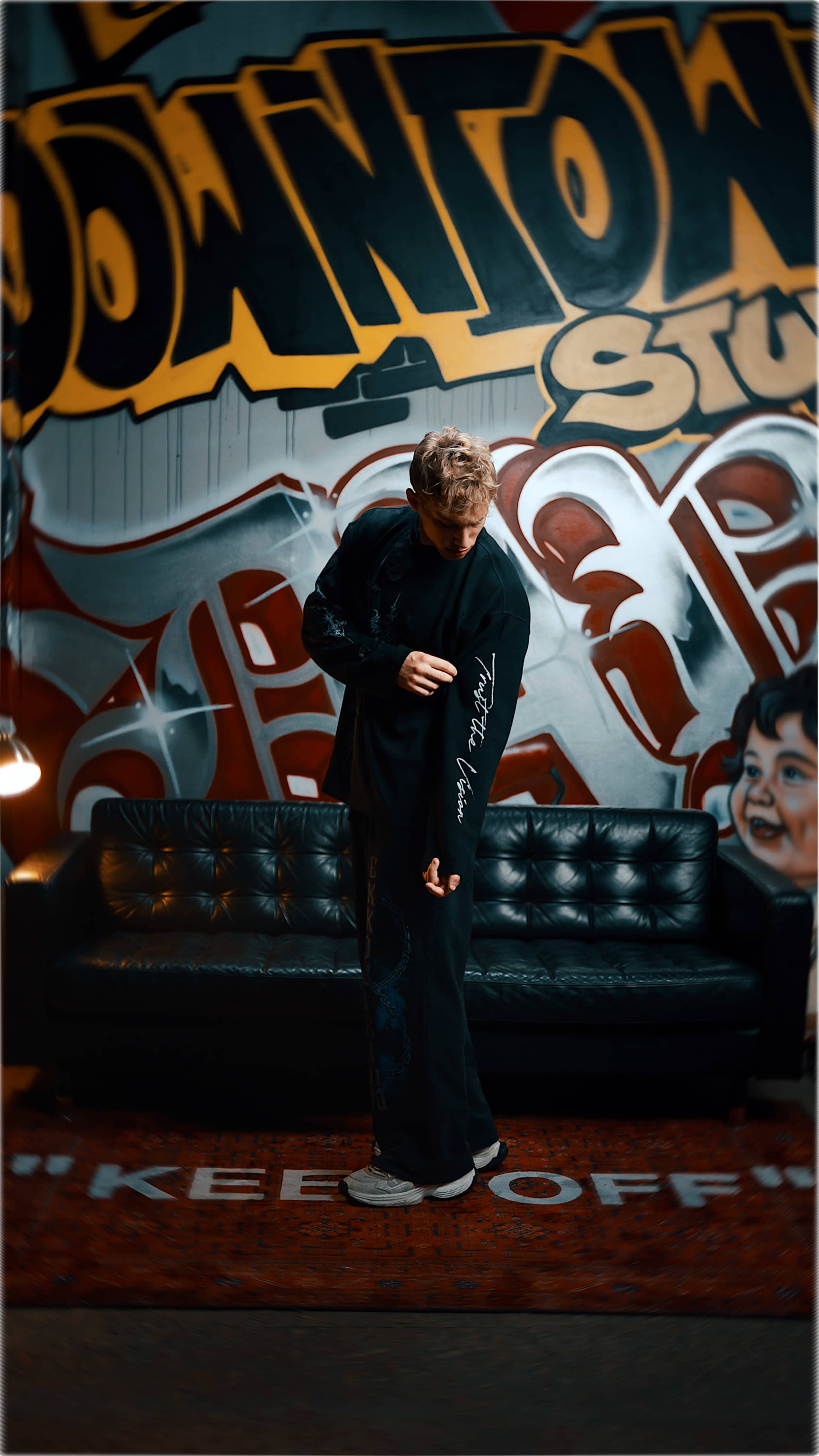 A person in casual attire stands in front of a graffiti-covered wall, adjusting the sleeve of their shirt near a black couch.