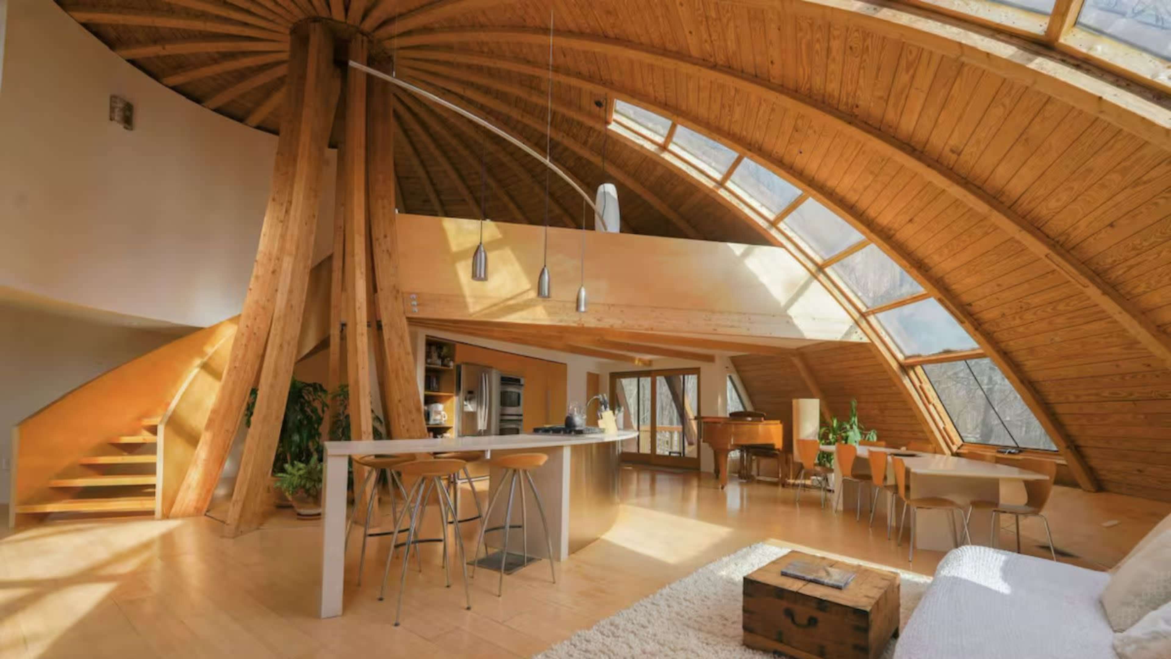 The image shows a modern, spacious interior of a home featuring a curved wooden structure with large windows and a mix of dining and living areas.