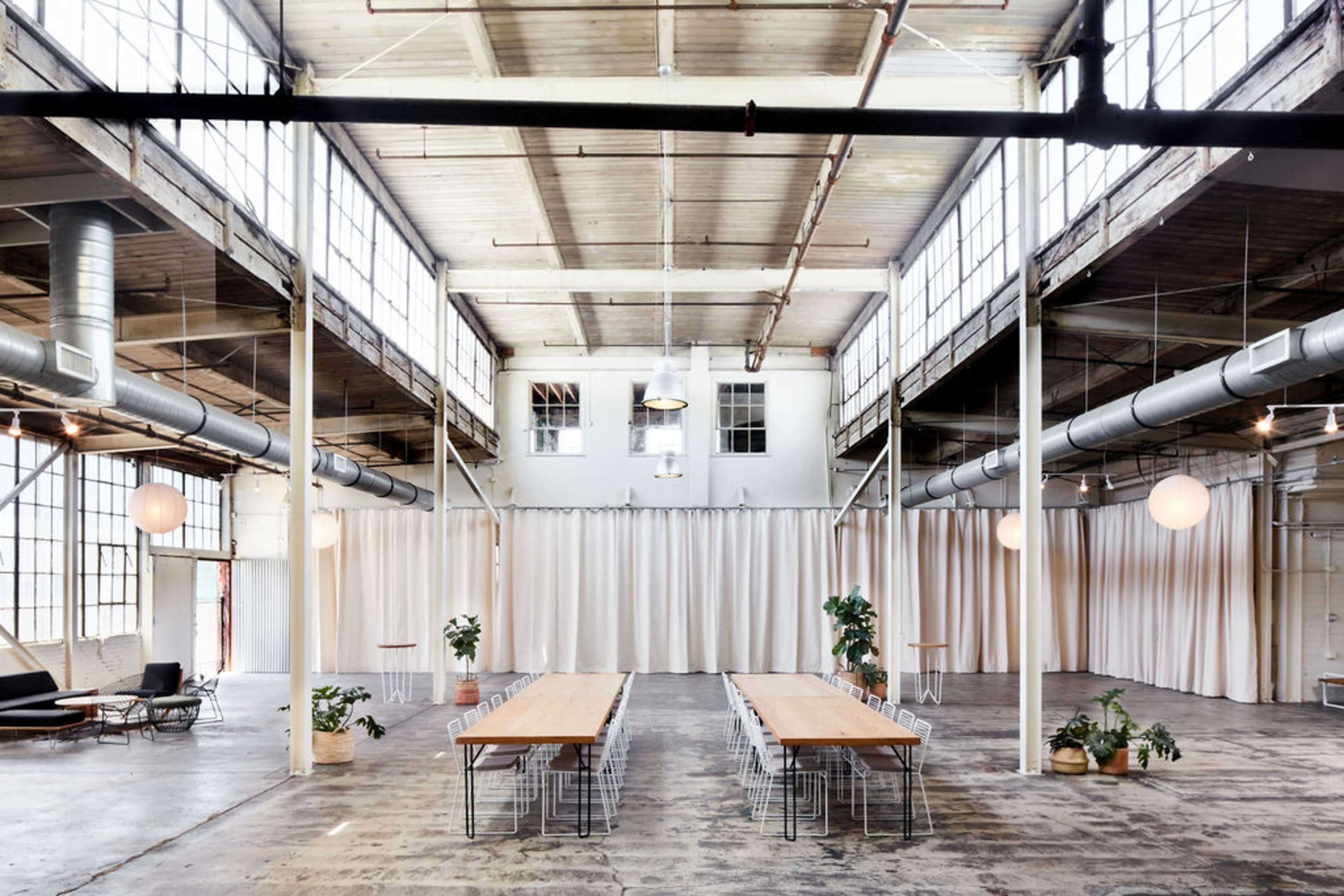A large, industrial-style room features multiple long tables arranged for gatherings, surrounded by potted plants and soft drapery.