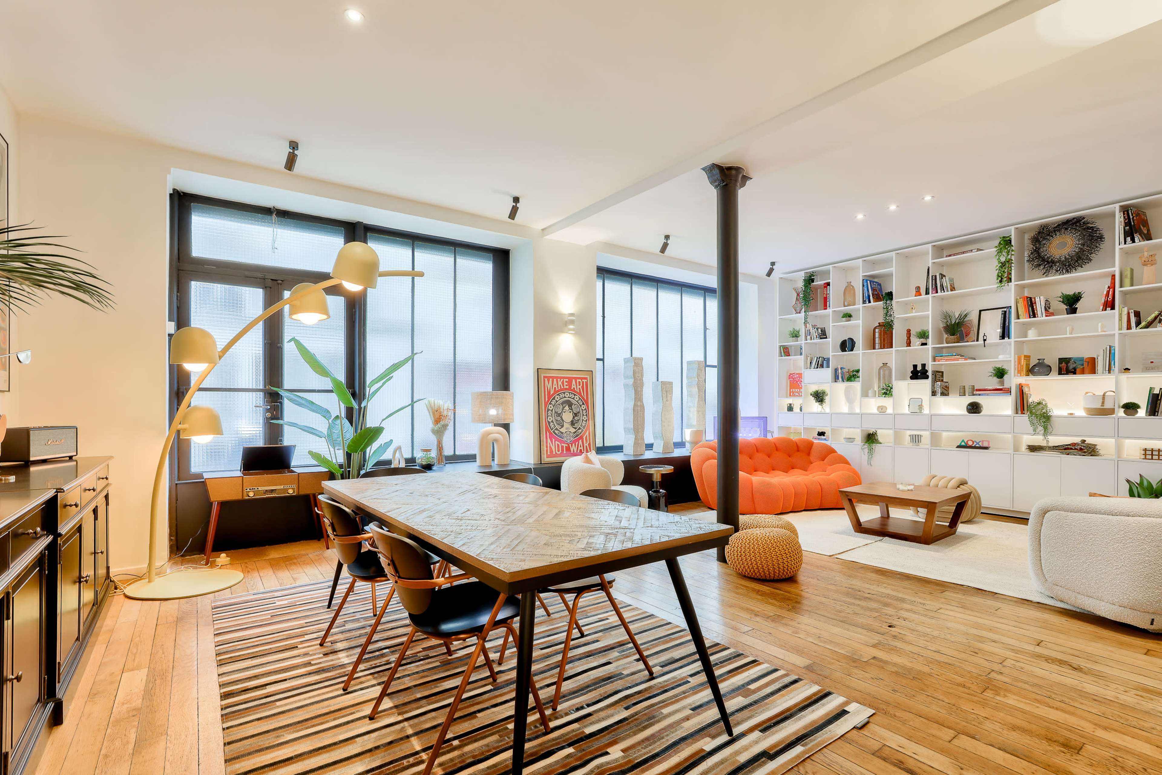 A modern living space features a large orange sofa, a dining table with black chairs, and shelves filled with decorative items, illuminated by natural light from large windows.