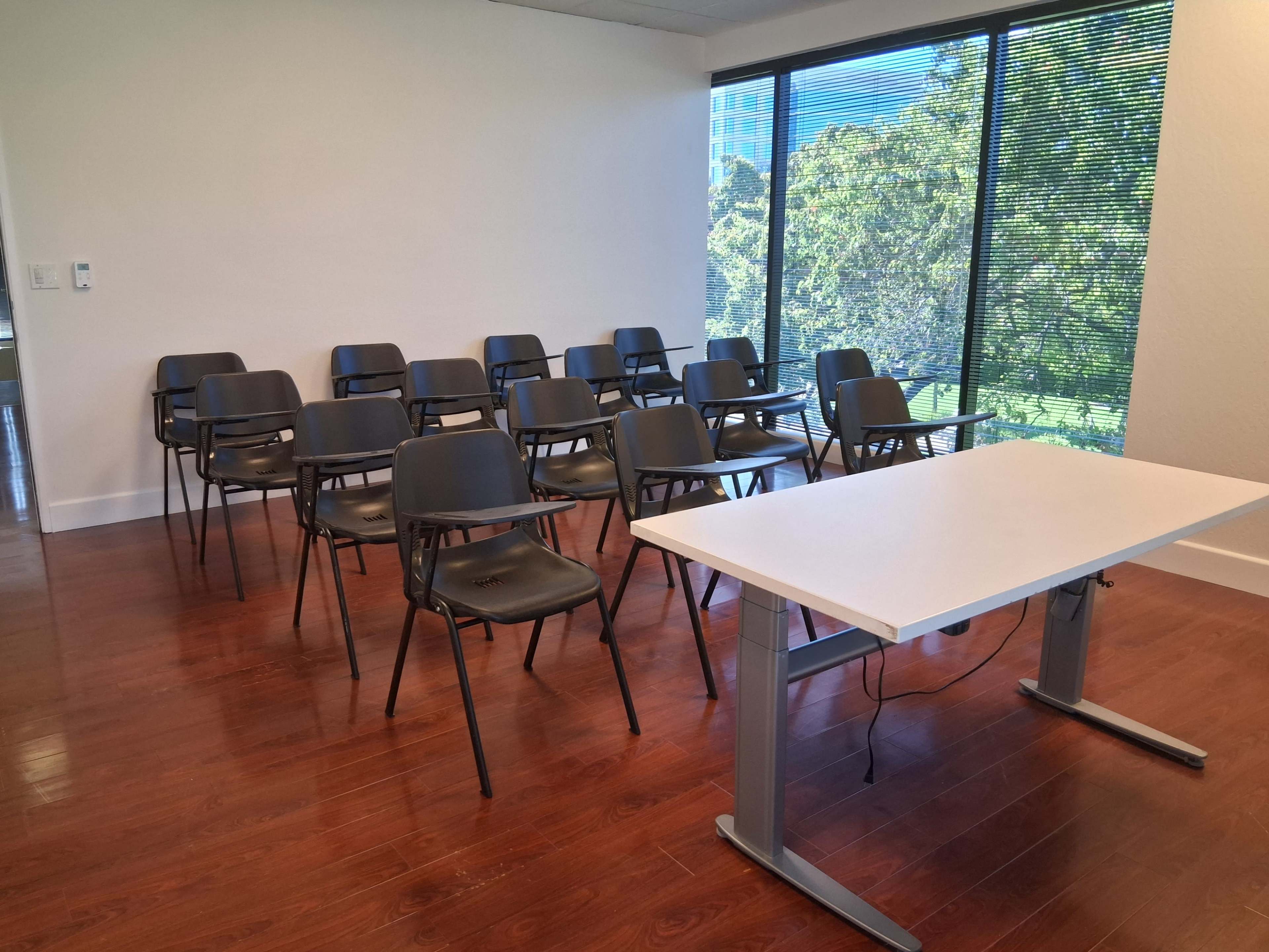 The image shows a conference room with a row of black chairs facing a white table, situated by large windows that overlook greenery outside.