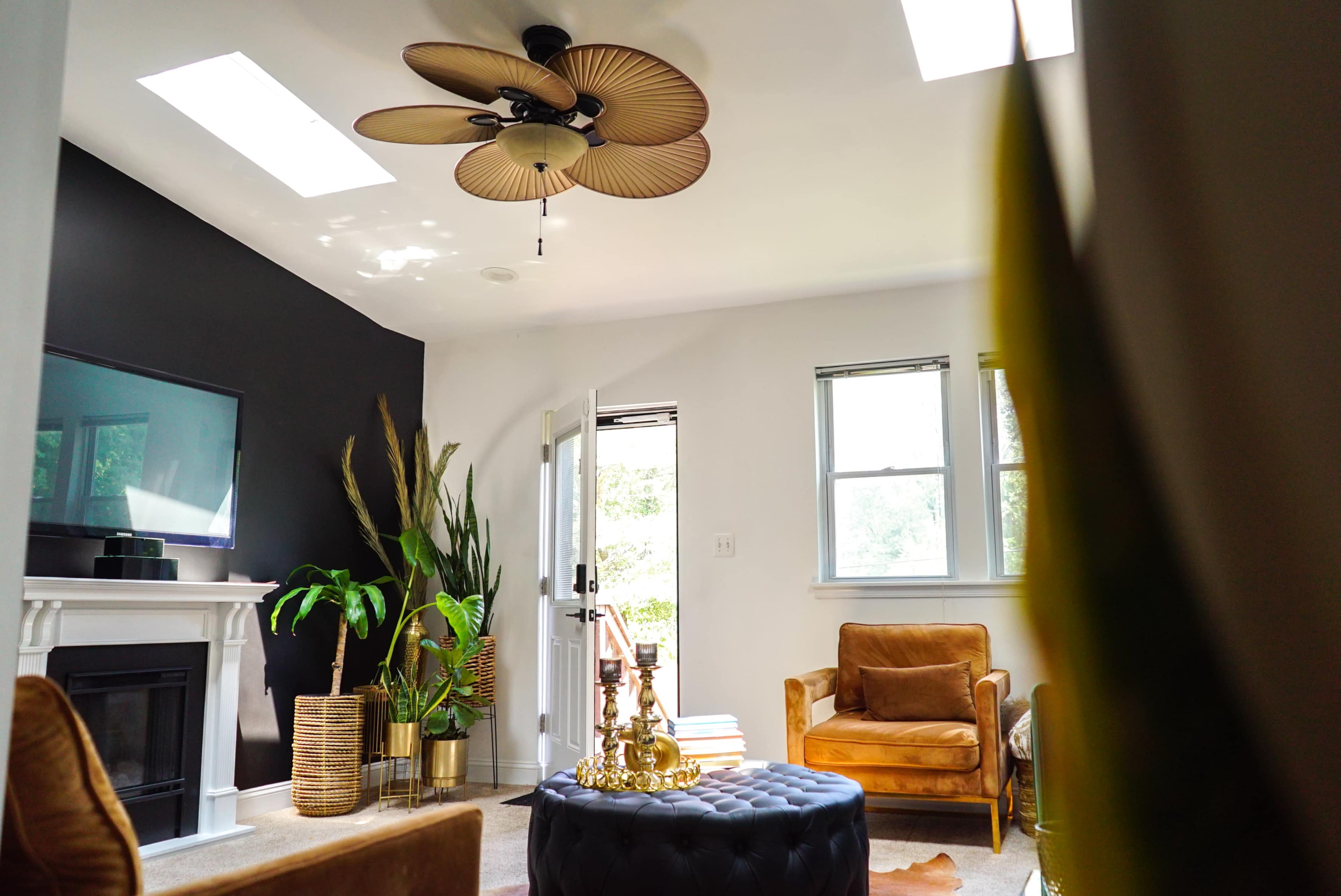 A contemporary living room features a black accent wall, a ceiling fan, potted plants, a brown armchair, and a round ottoman.