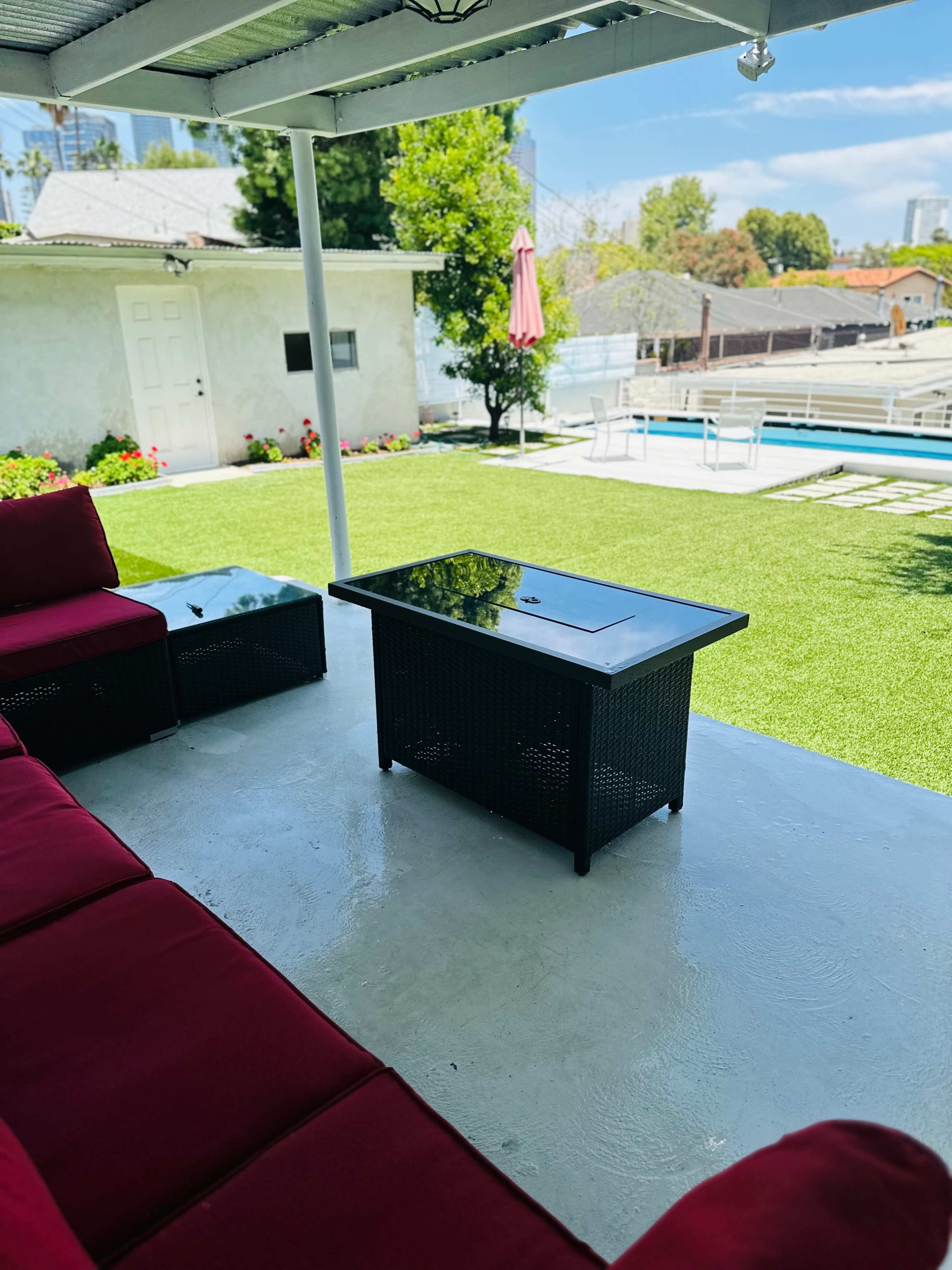 A covered outdoor seating area features red couches and a glass-top coffee table overlooking a green lawn and a swimming pool.