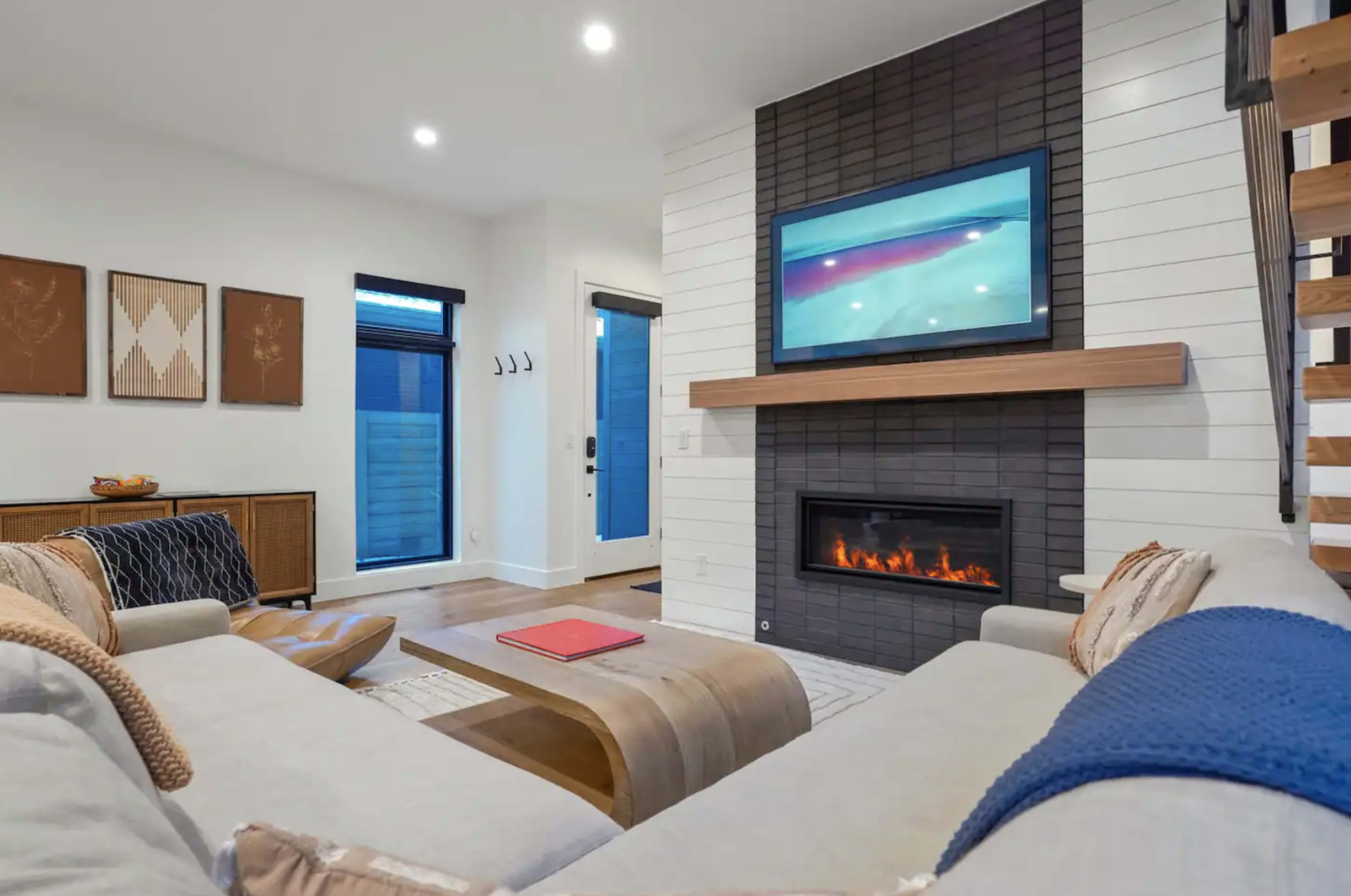 A modern living room features a cozy seating area, a fireplace beneath a mounted television, and large windows allowing natural light.