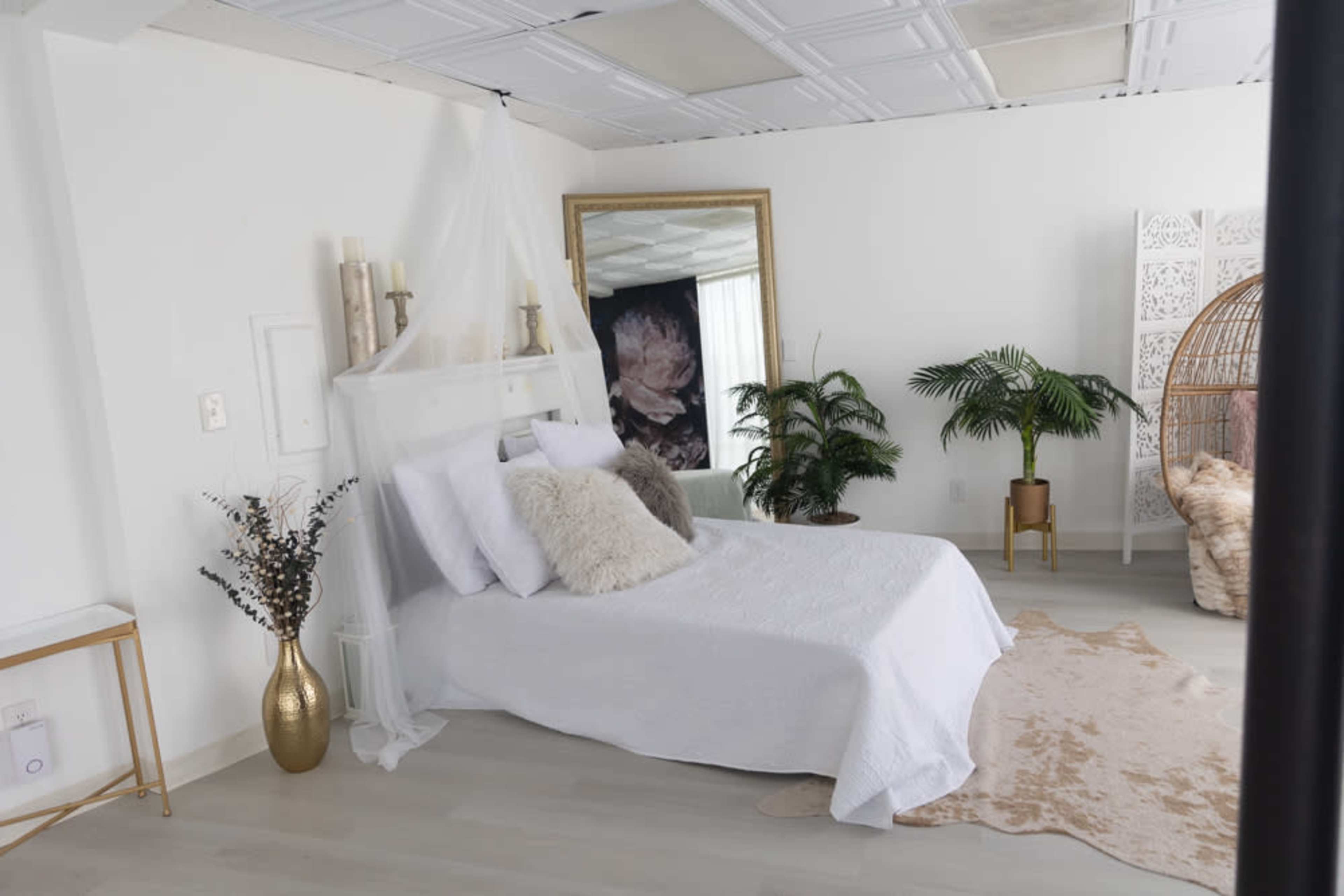 A bright, minimalist bedroom featuring a white bed with decorative pillows, a large mirror, plants, and a cozy seating area.