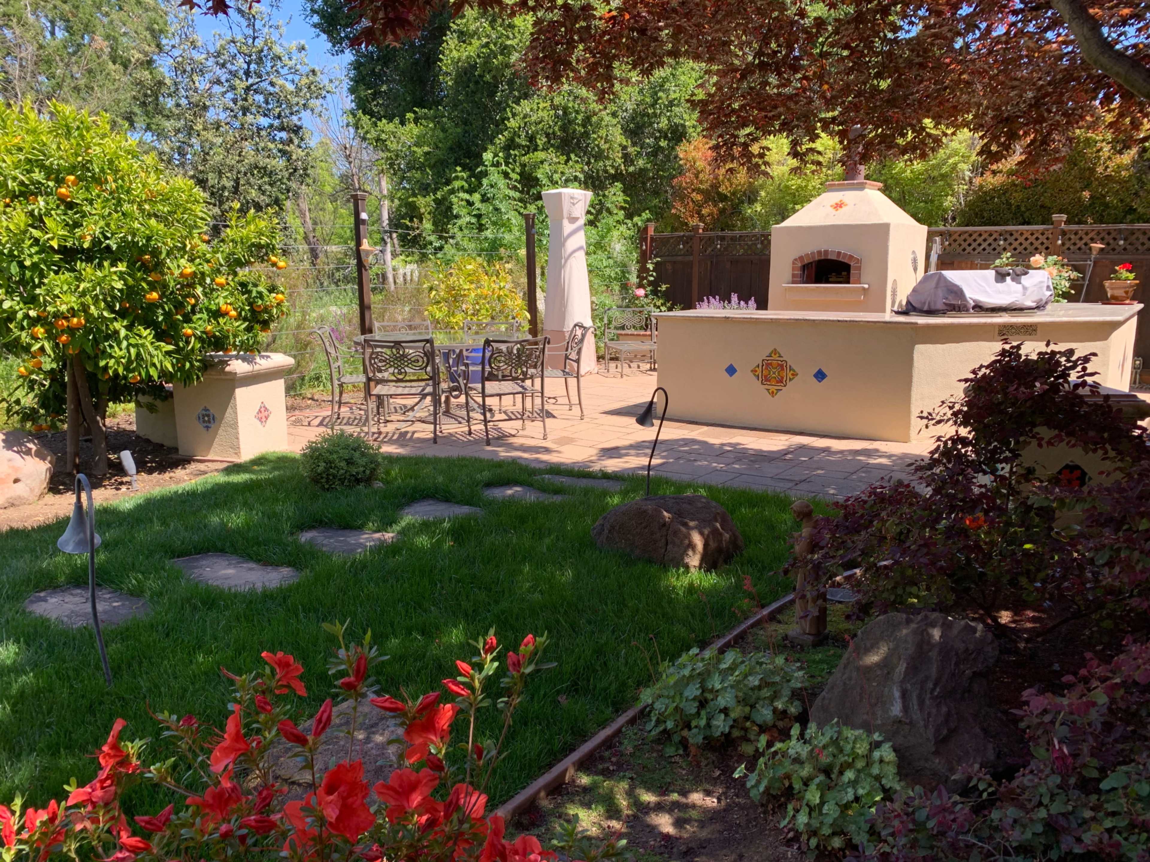 A landscaped backyard with a stone patio, an outdoor kitchen, a dining set, and various plants and trees.