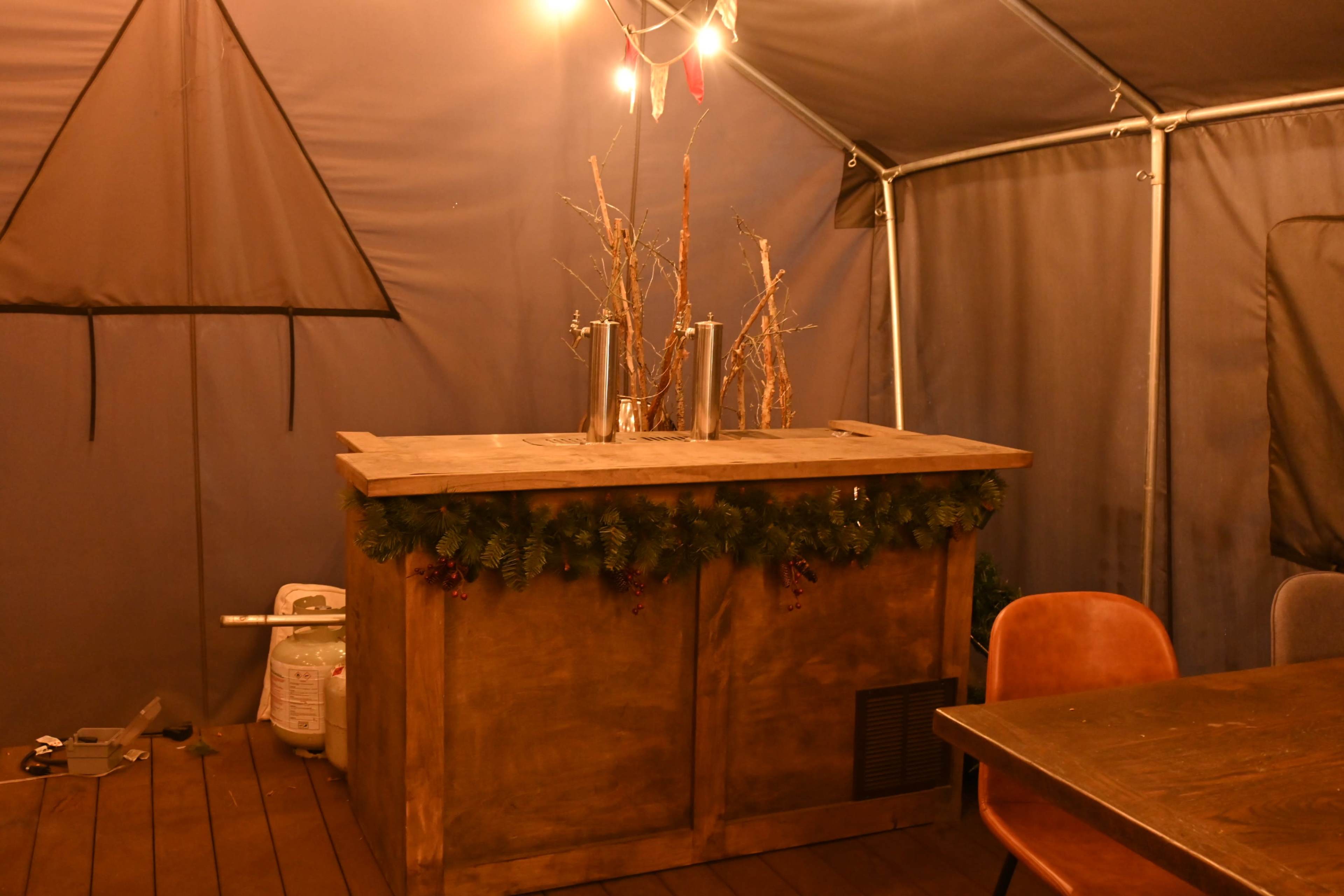 The image shows a wooden bar counter decorated with greenery and a few decorative branches, set in a tent-like enclosure with dim lighting.
