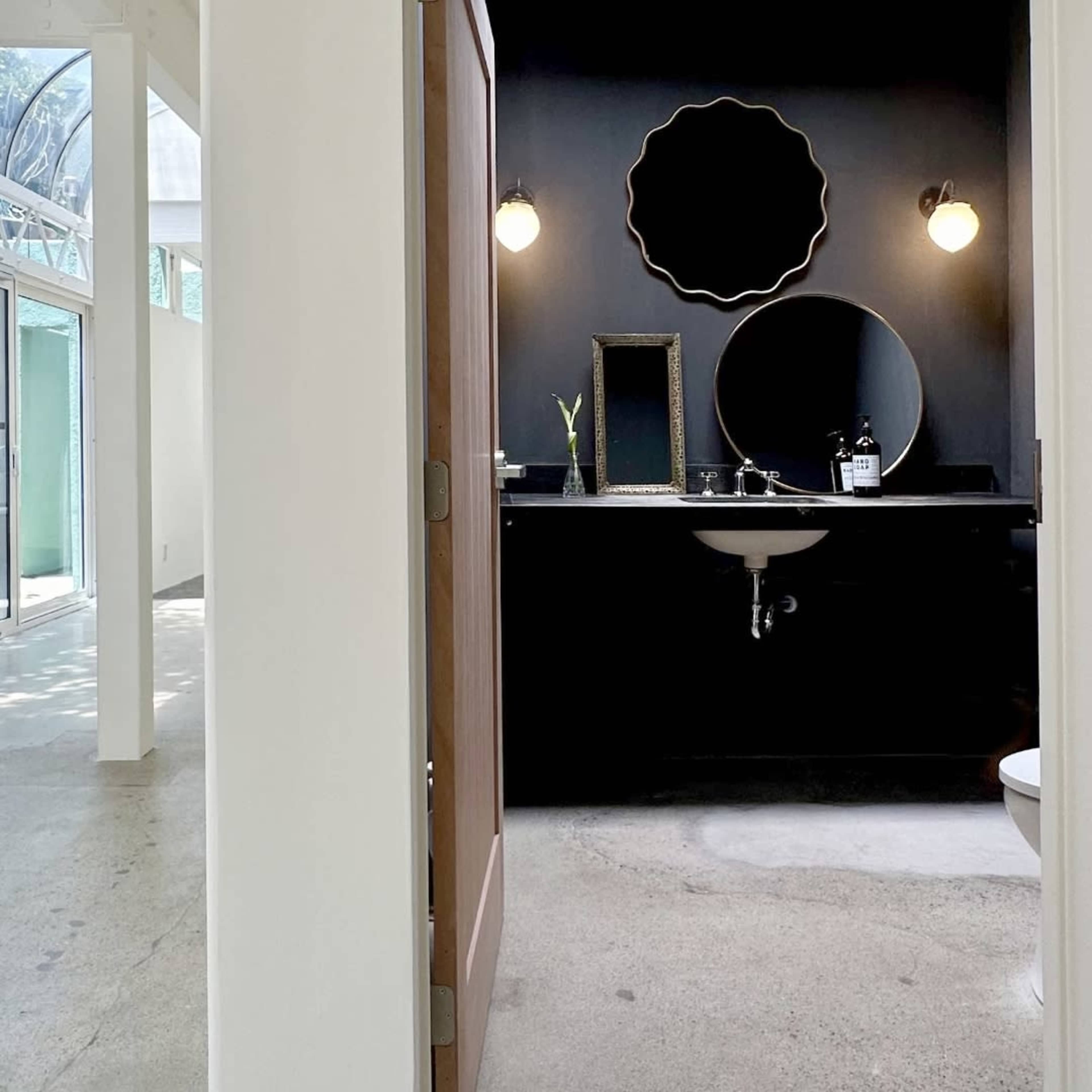 A modern bathroom features a dark wall with two round mirrors and a sink, adjacent to a light-filled space with large windows.