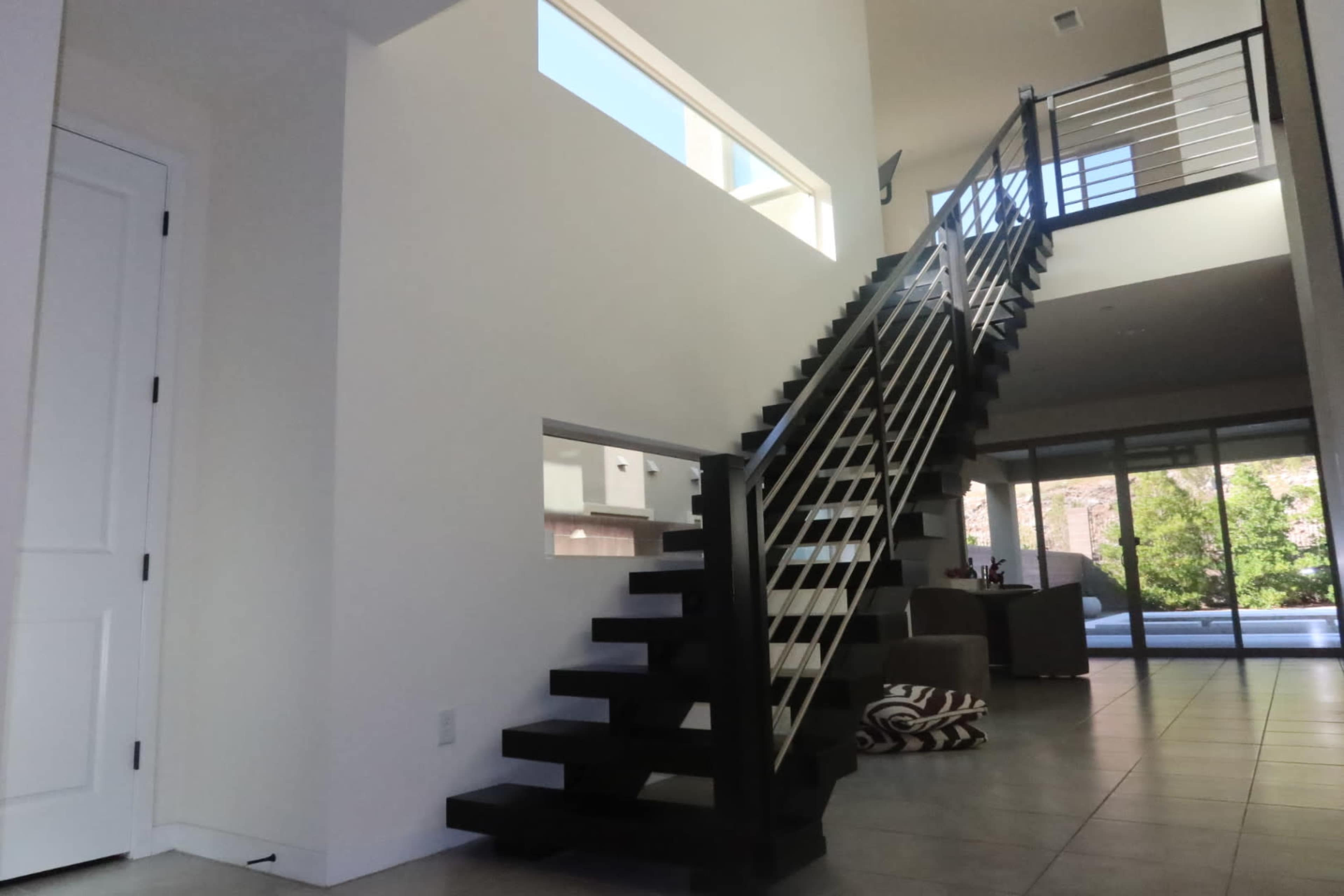 A modern interior features a staircase with black wooden steps and metal railings, leading to a bright upper level, with a view of large windows and a living area in the background.
